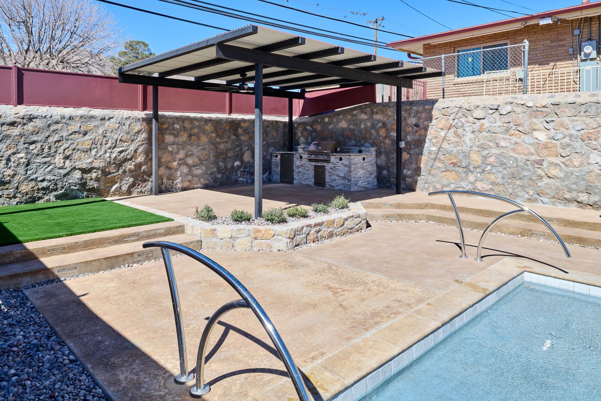 Outdoor patio with a covered grilling station, stone walls, tiered concrete levels, synthetic grass, and a swimming pool.