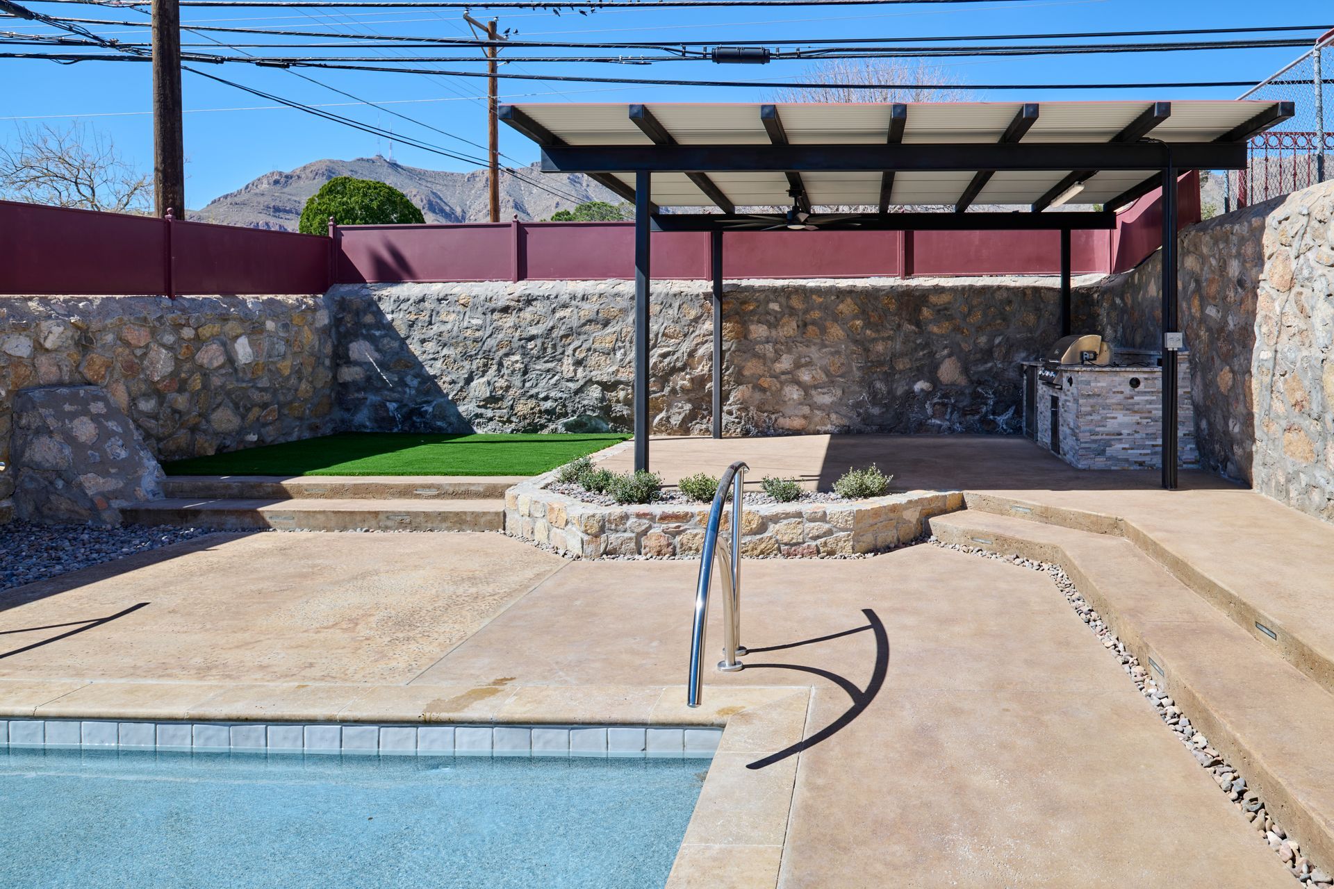 A swimming pool area with concrete patio, stone retaining walls, a covered pergola, and small patch of green grass.