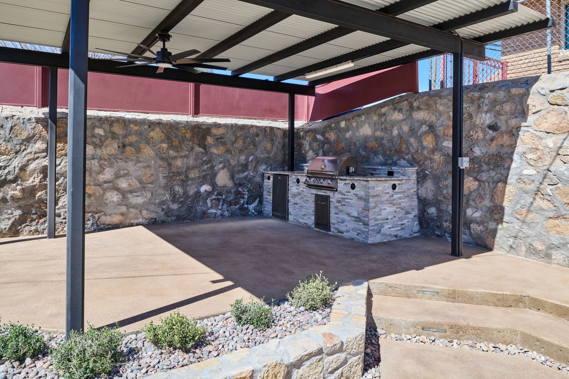 An outdoor covered patio area with stone walls, a built-in stone grill station, a ceiling fan, and landscaped planters.