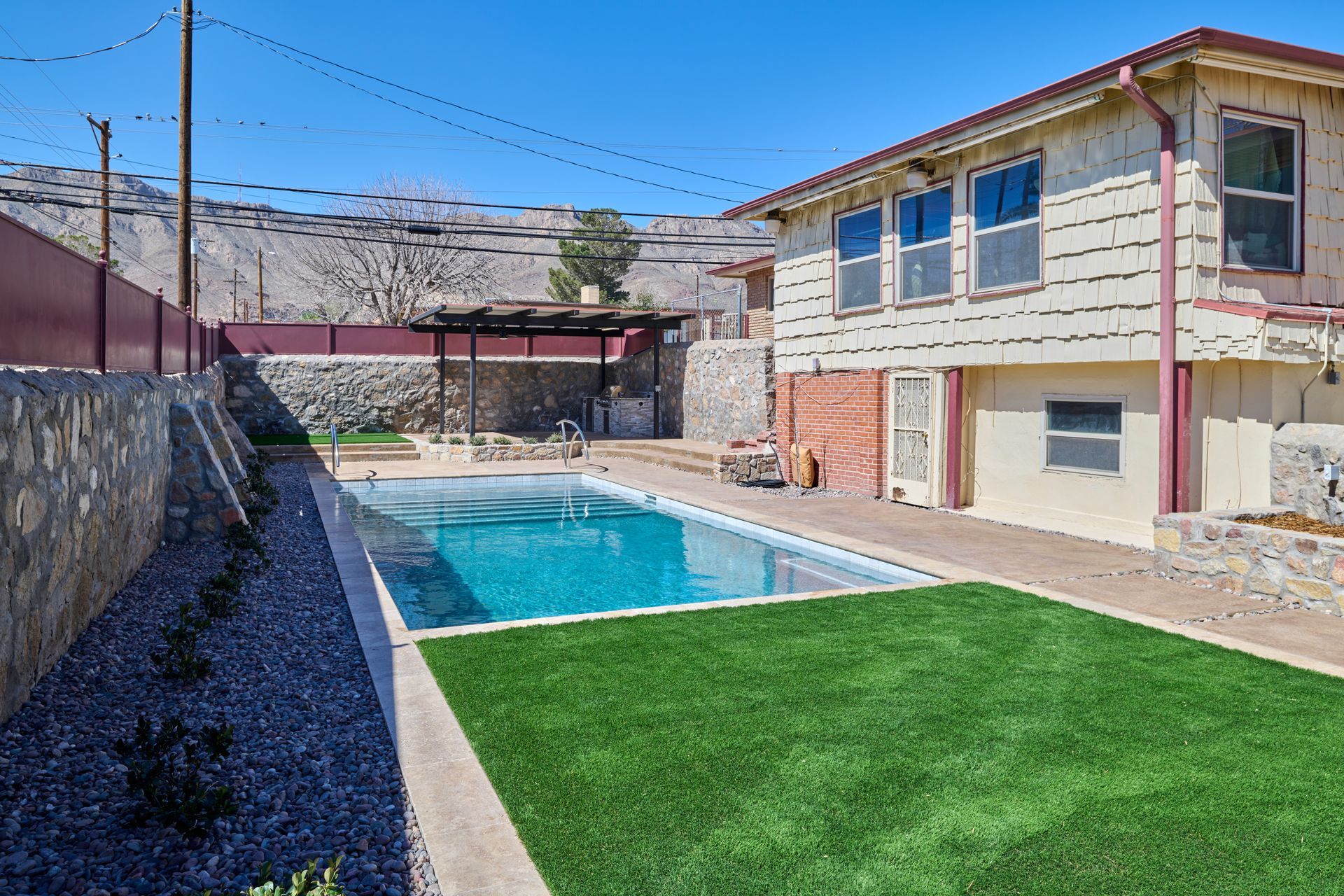 A backyard features a rectangular swimming pool, green artificial turf, a stone retaining wall, and a beige house.