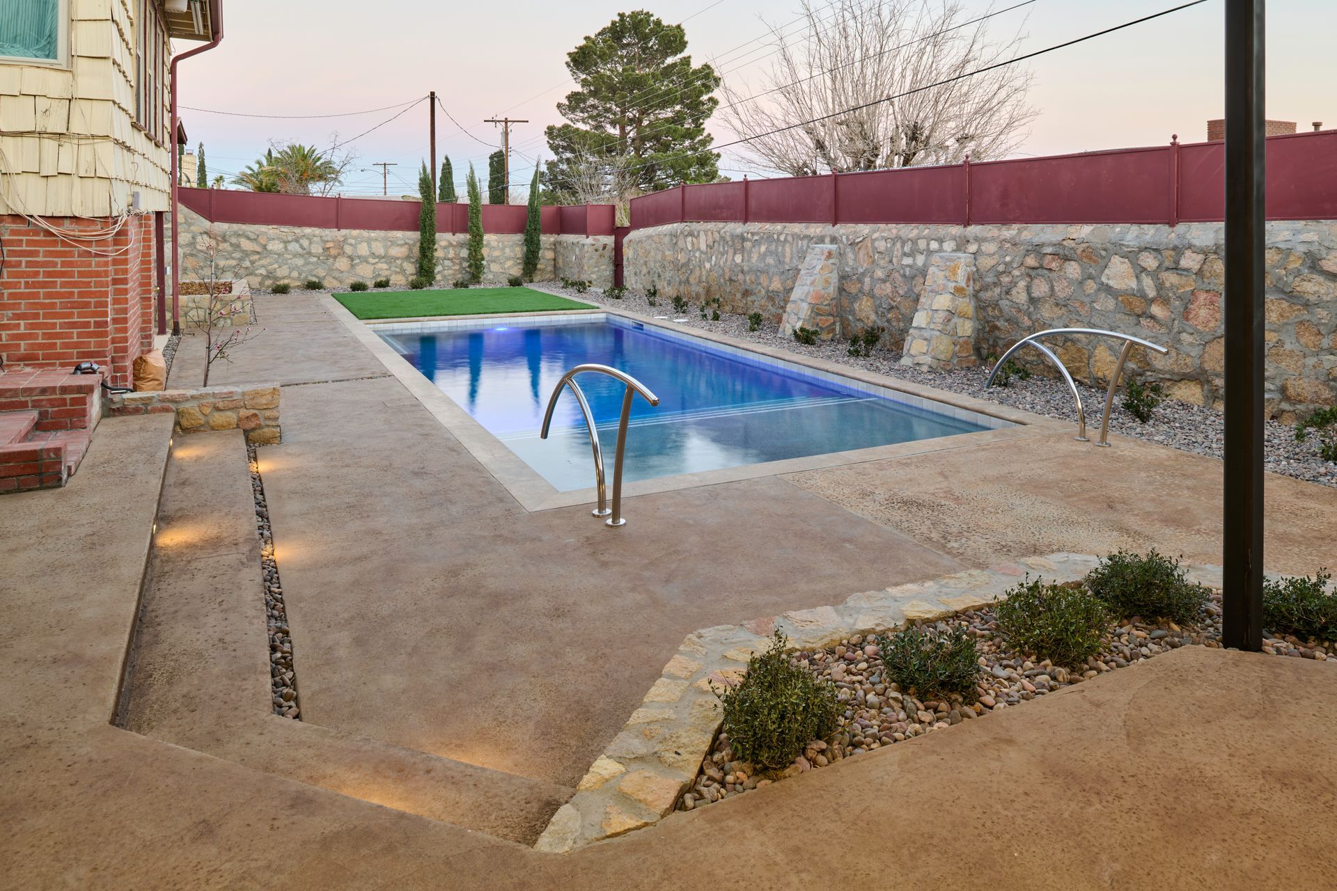 A backyard features a rectangular swimming pool surrounded by a textured patio, stone walls, and desert landscaping.