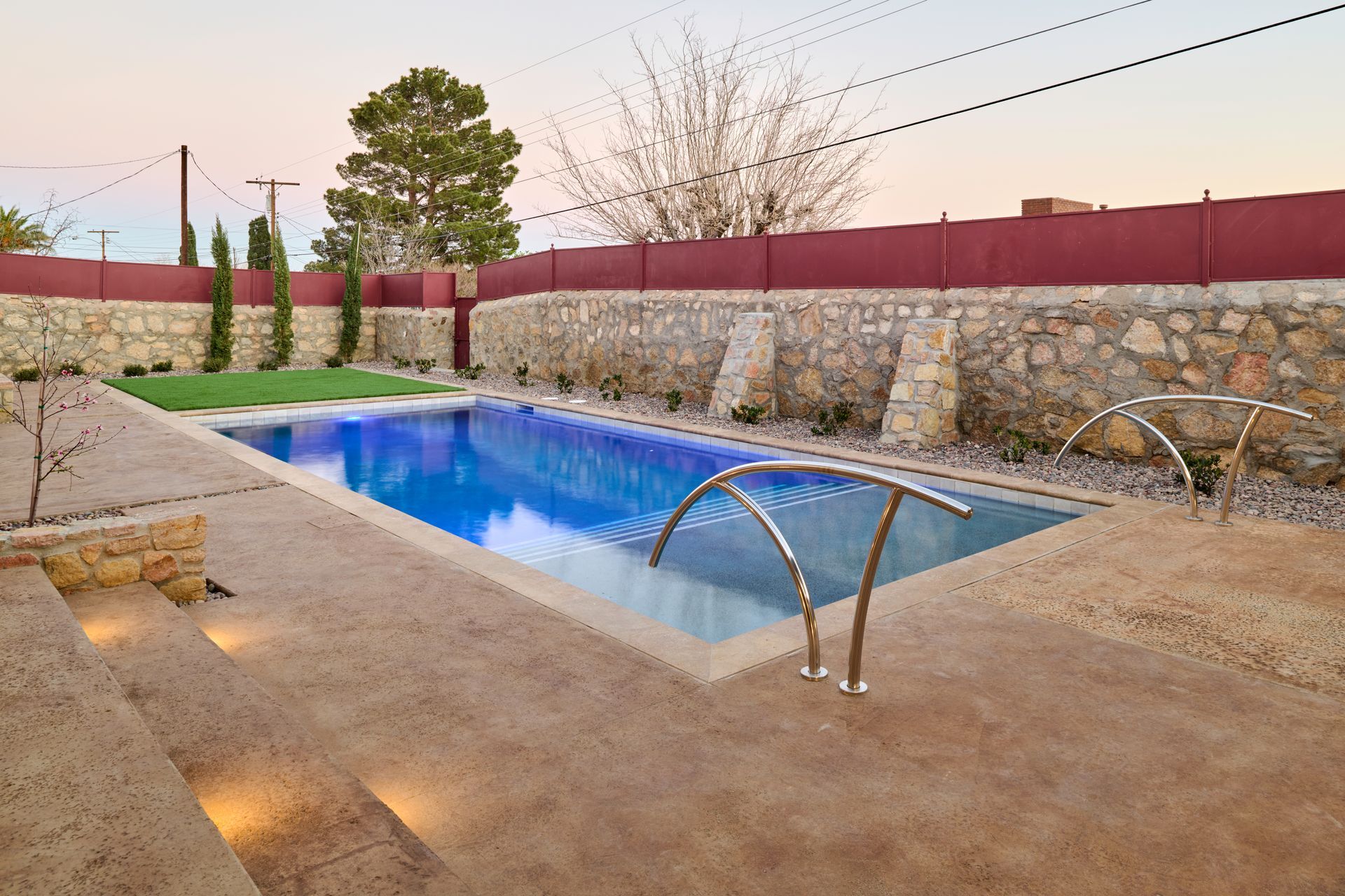 A blue rectangular swimming pool with steel handrails, surrounded by stone walls and a textured patio at twilight.