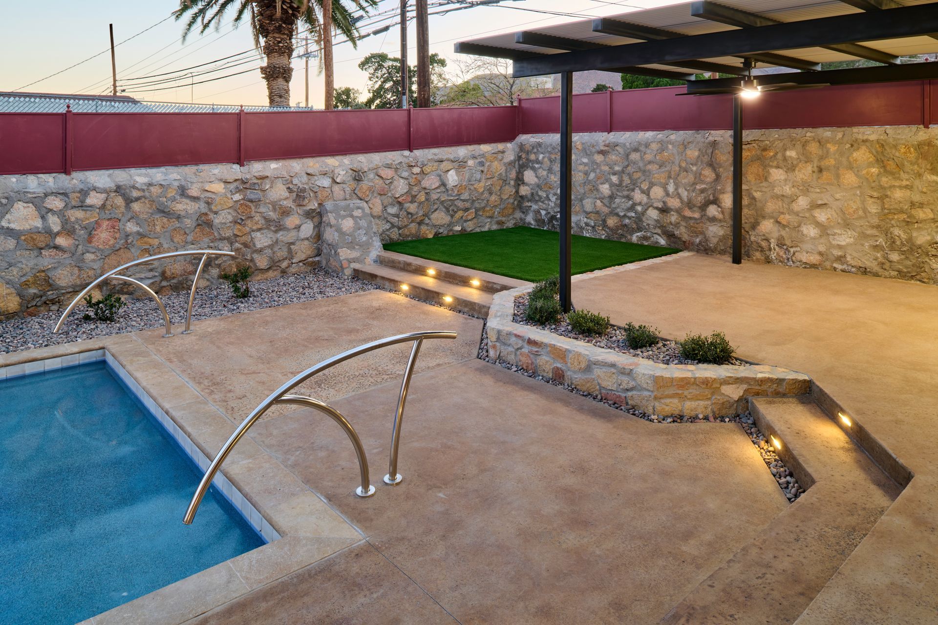 A modern backyard with a pool, stone walls, a covered patio area, and integrated landscape lighting at dusk.