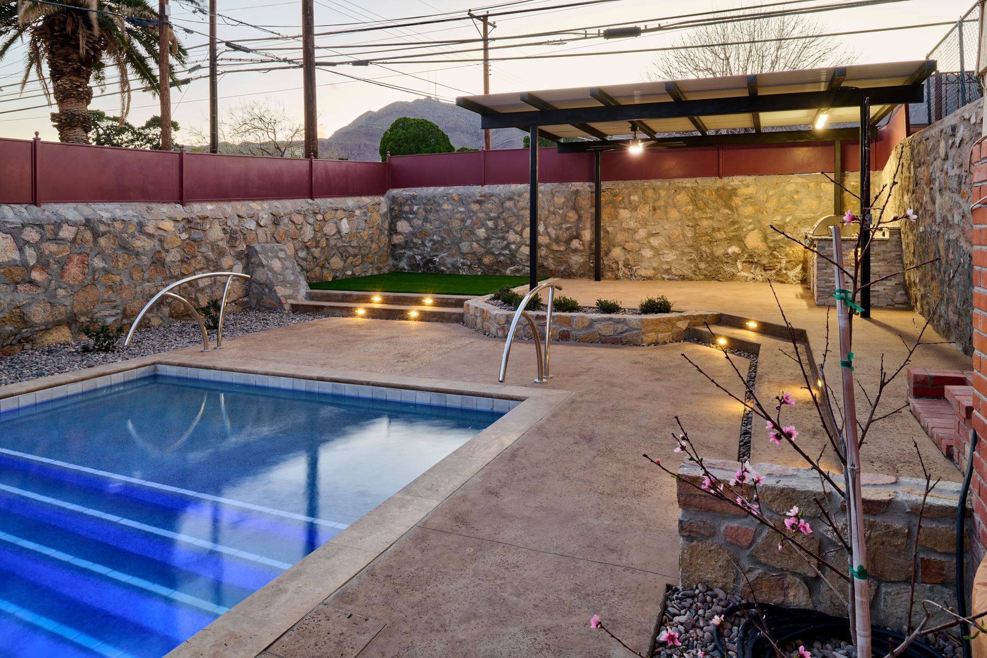 A backyard area featuring a rectangular pool, stone walls, a covered patio structure, and outdoor lighting at dusk.