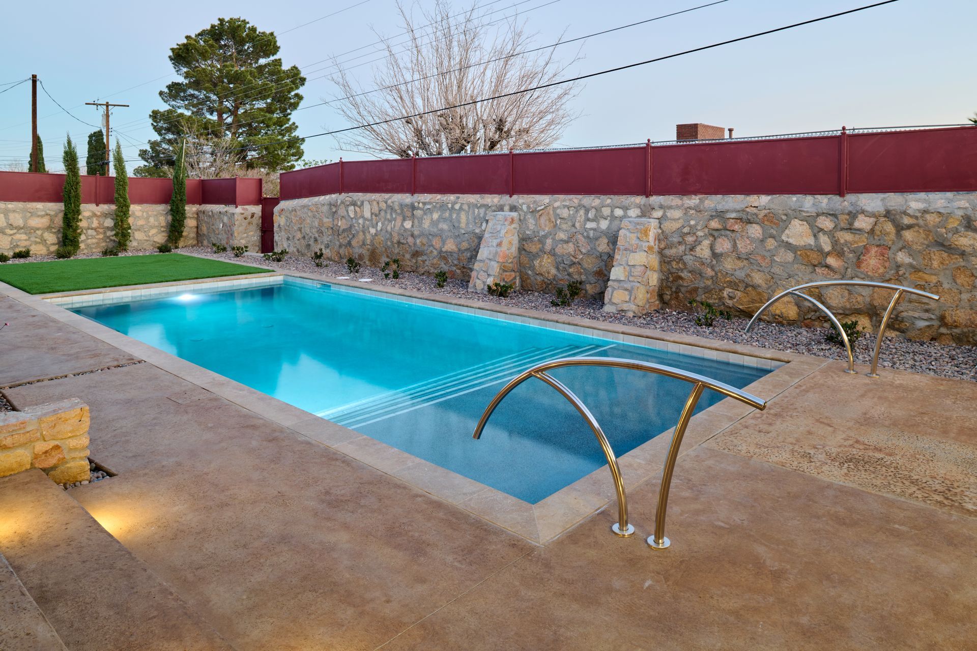 A rectangular swimming pool with blue water sits in a patio area with a stone wall, red fence, and grass.