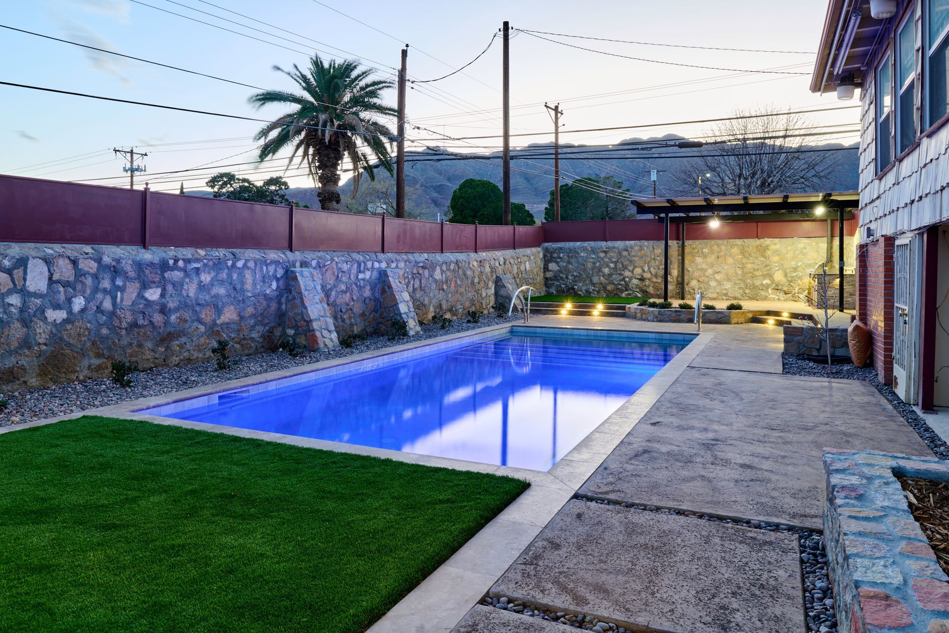 A rectangular swimming pool with bright blue water sits in a courtyard with a stone wall, artificial lawn, and patio.