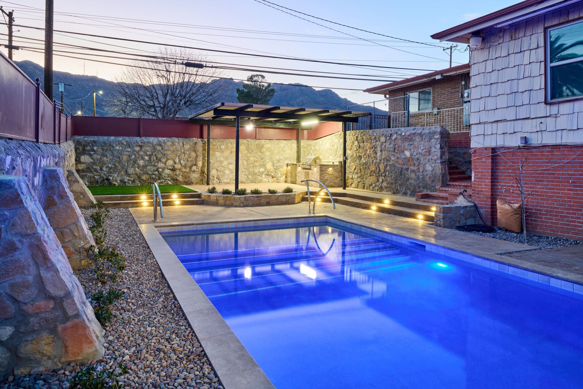 An illuminated backyard swimming pool at dusk, with a stone-walled patio and a covered outdoor structure in the back.