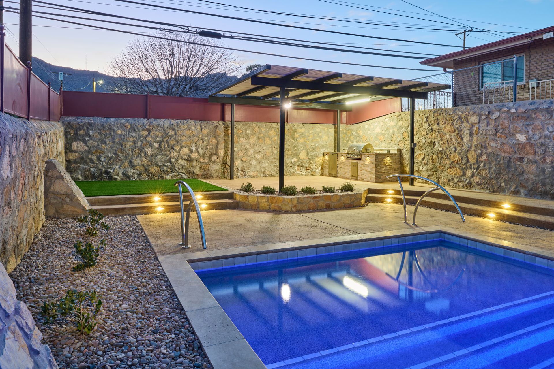 A swimming pool and patio area at dusk, featuring a covered outdoor kitchen, stone walls, and landscape lighting.