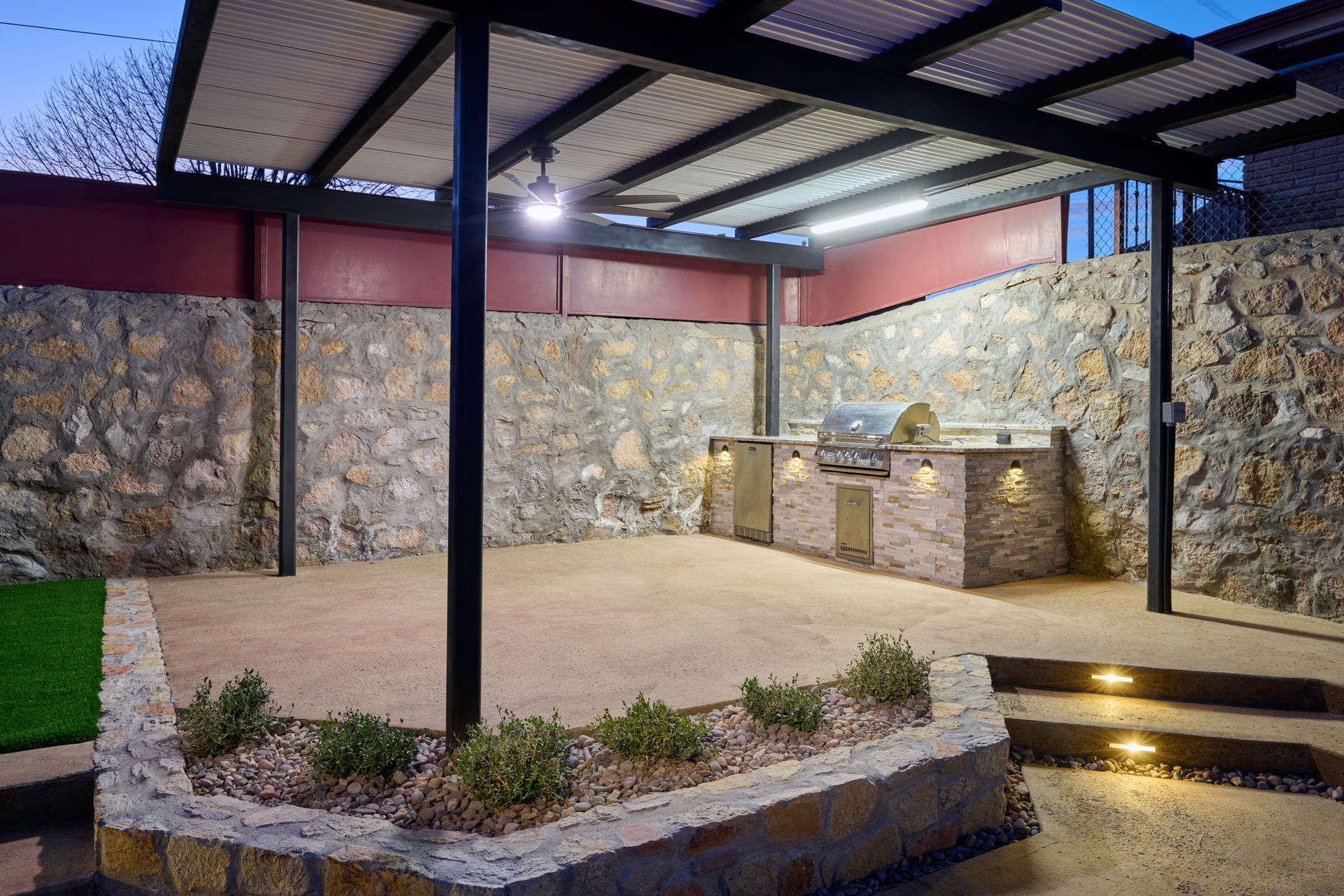 A covered outdoor patio featuring a stone grill station, rustic rock walls, and lighting at dusk.