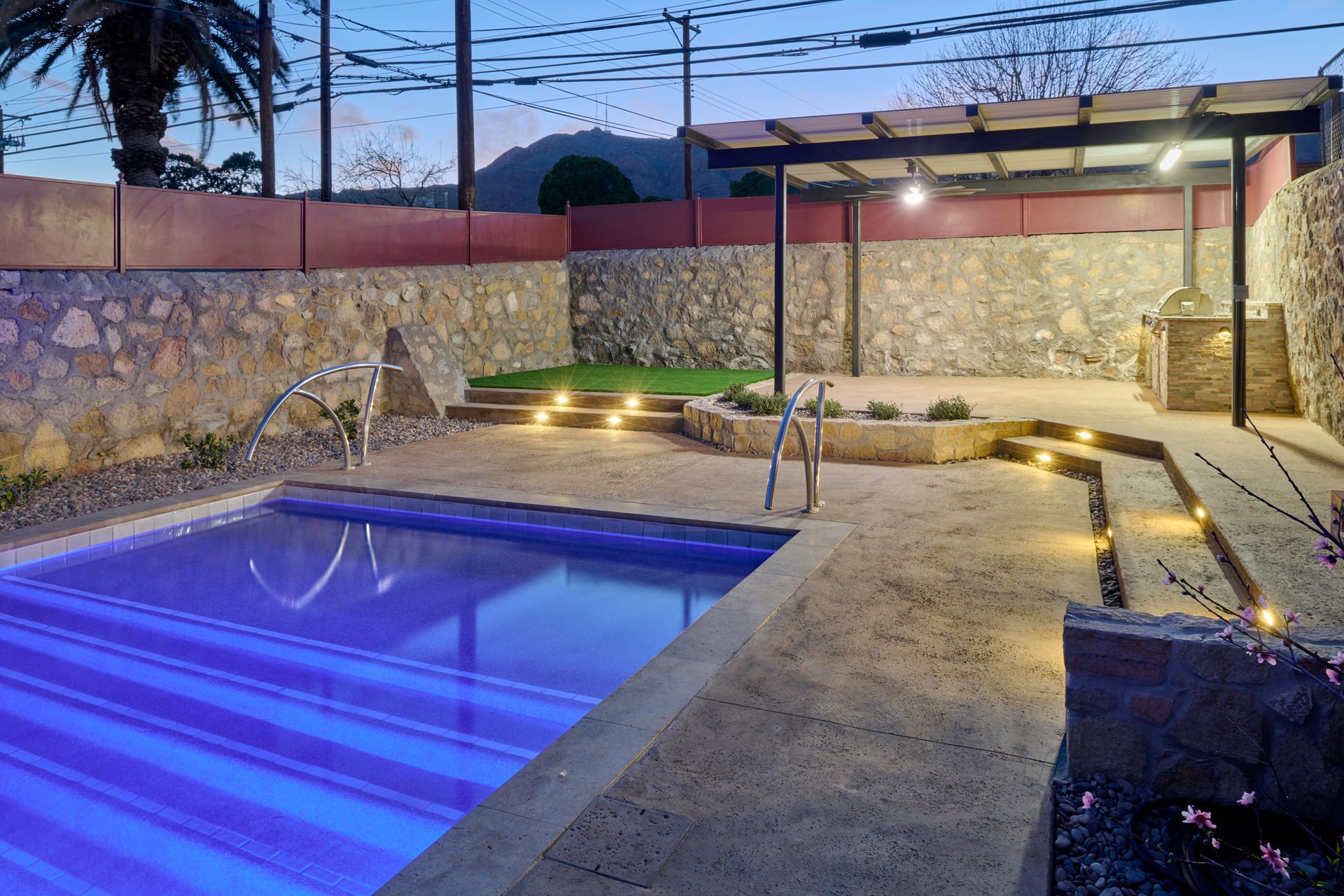 Outdoor pool with blue water and lit steps at dusk, featuring a stone wall, pergola, and gravel patio area.