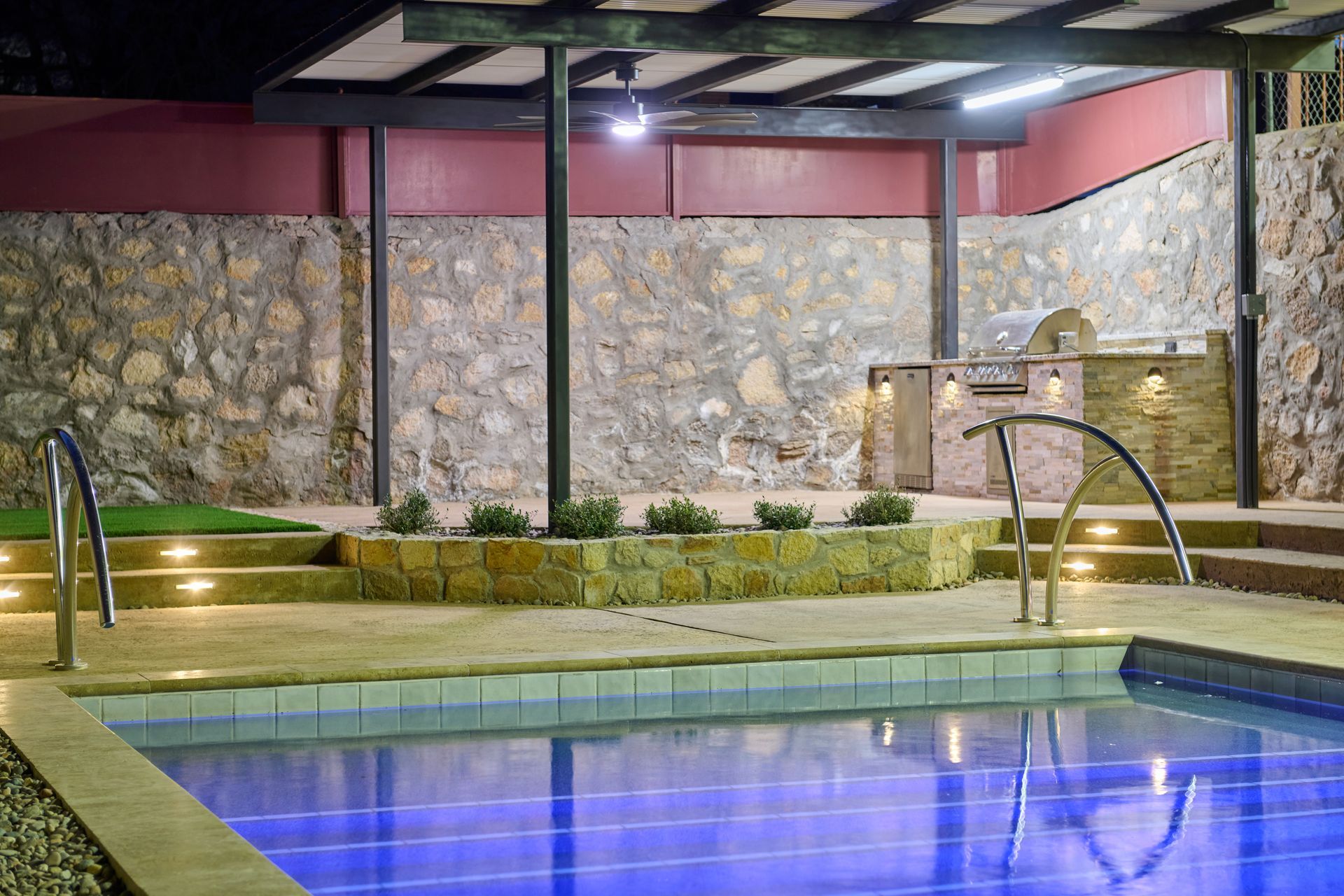 Illuminated swimming pool at night with a stone wall background, covered patio area, and built-in outdoor grill.