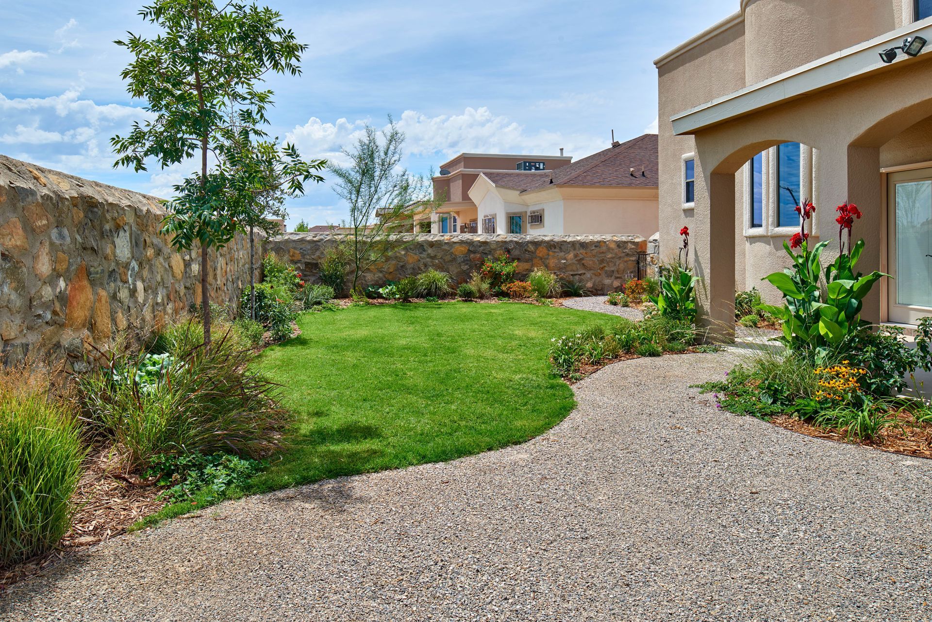 A gravel path curves towards a green lawn with plants, bordered by a stone wall and a beige house with red flowers.