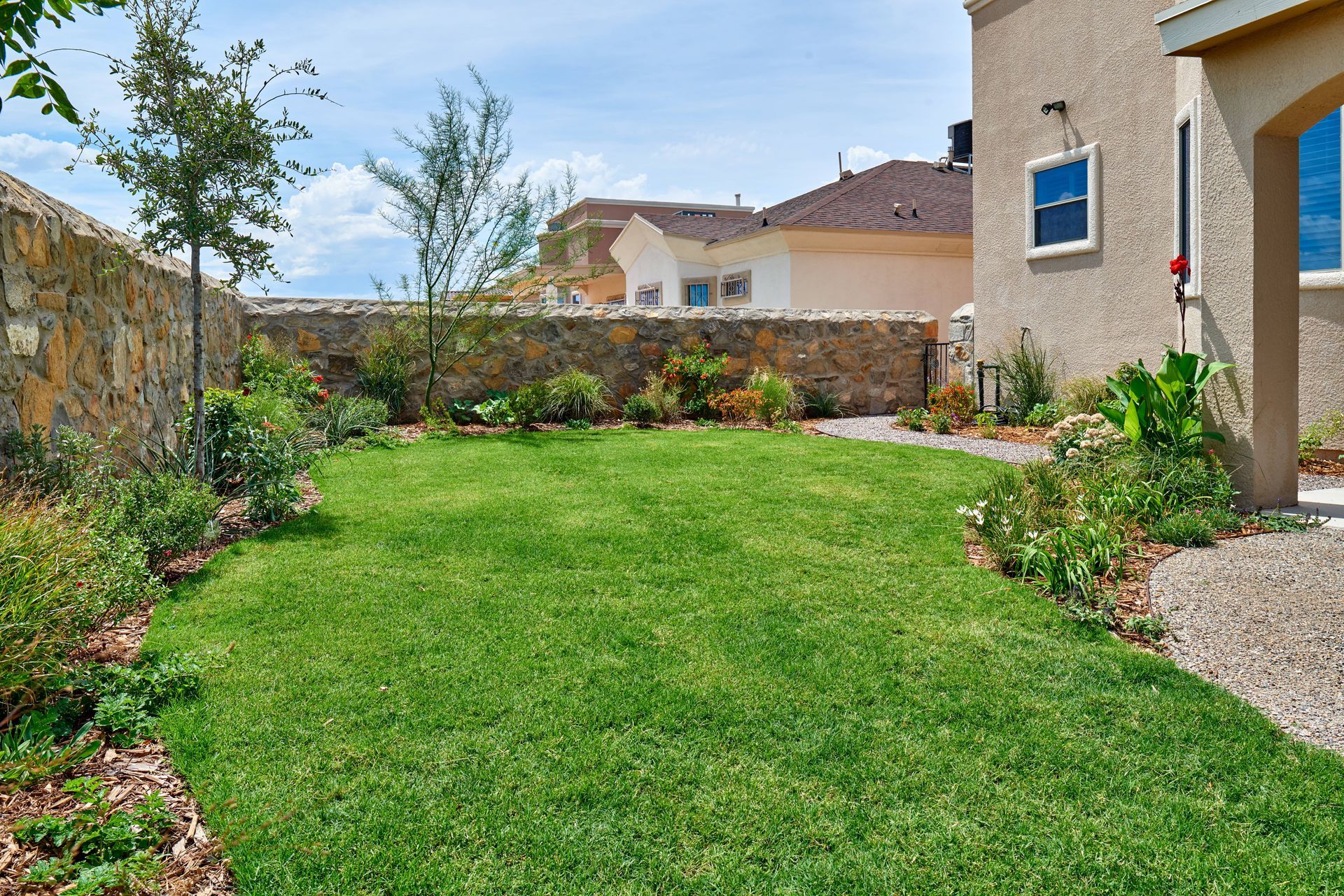 Lush green lawn in a backyard surrounded by stone walls and flowering plants under a sunny sky.