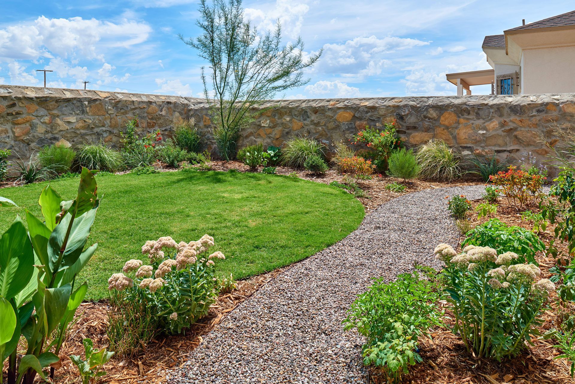 A garden with a grassy patch, mulch path, and stone wall under a blue sky.