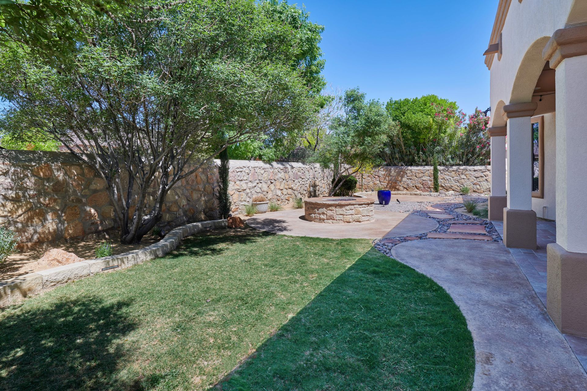 Backyard with green lawn, stone wall, tree, and patio. Sunny day.