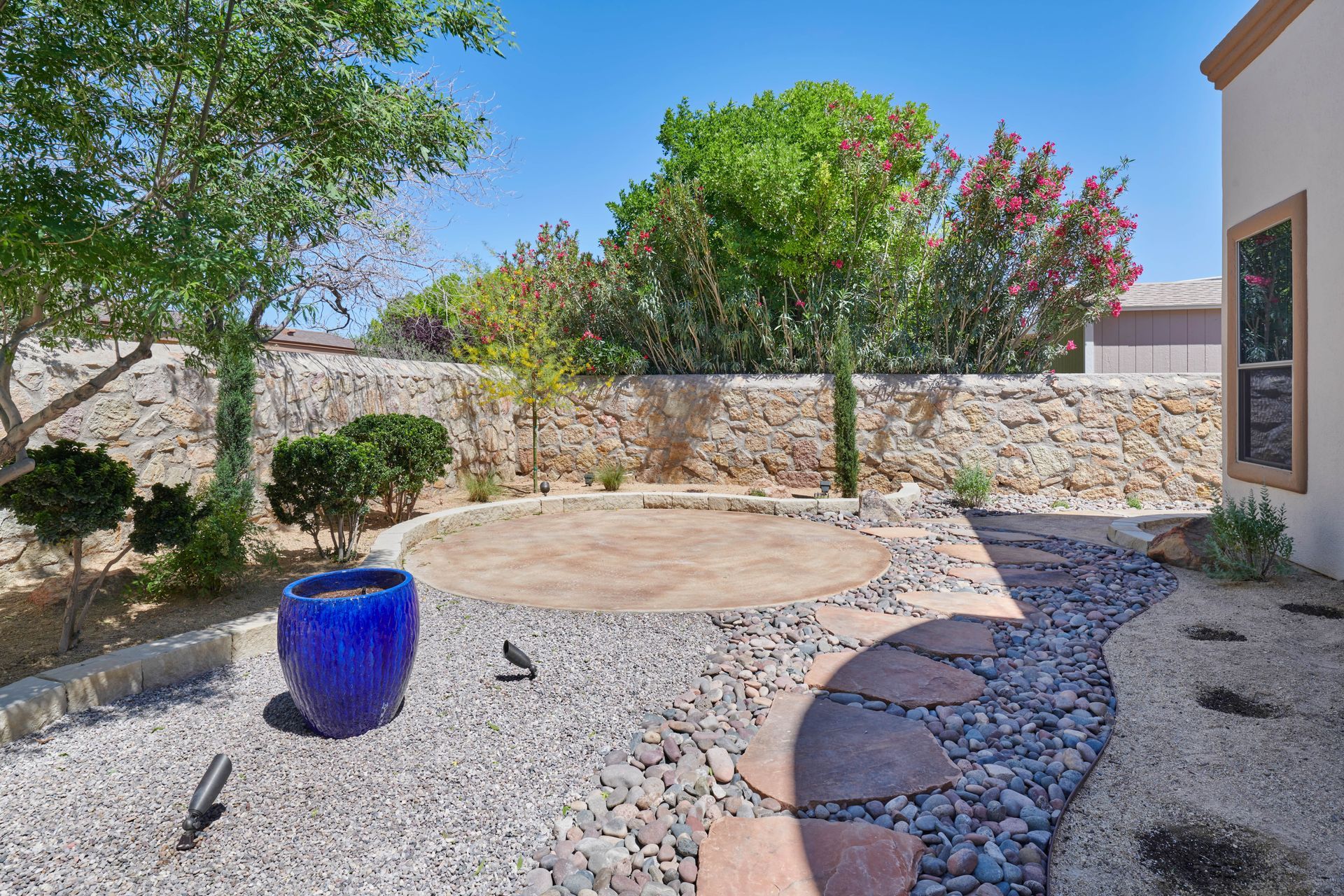 Backyard with stone wall, gravel, stepping stones, blue pot, and trees under a blue sky.