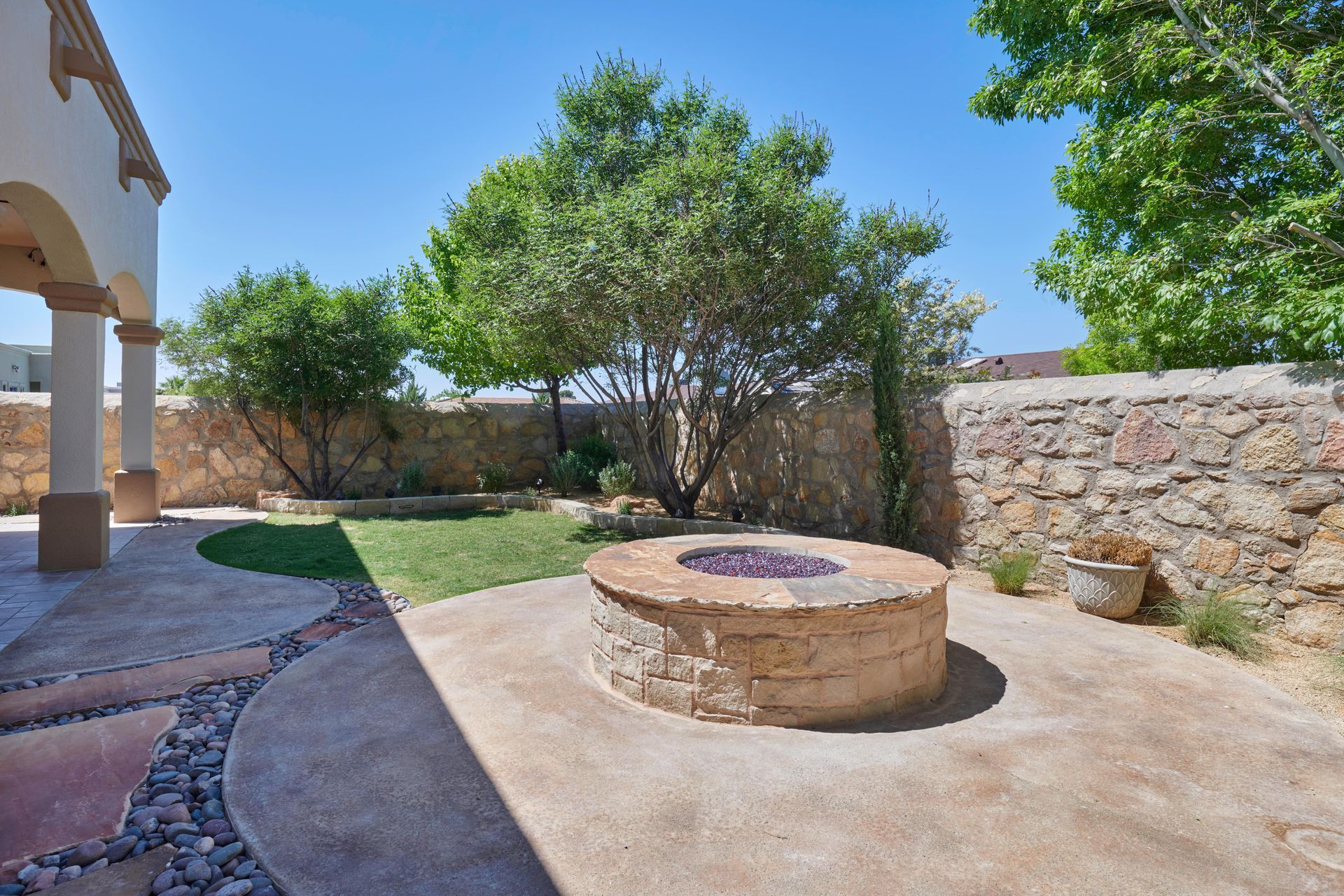 Stone fire pit on a patio with a lawn and stone walls under a blue sky.