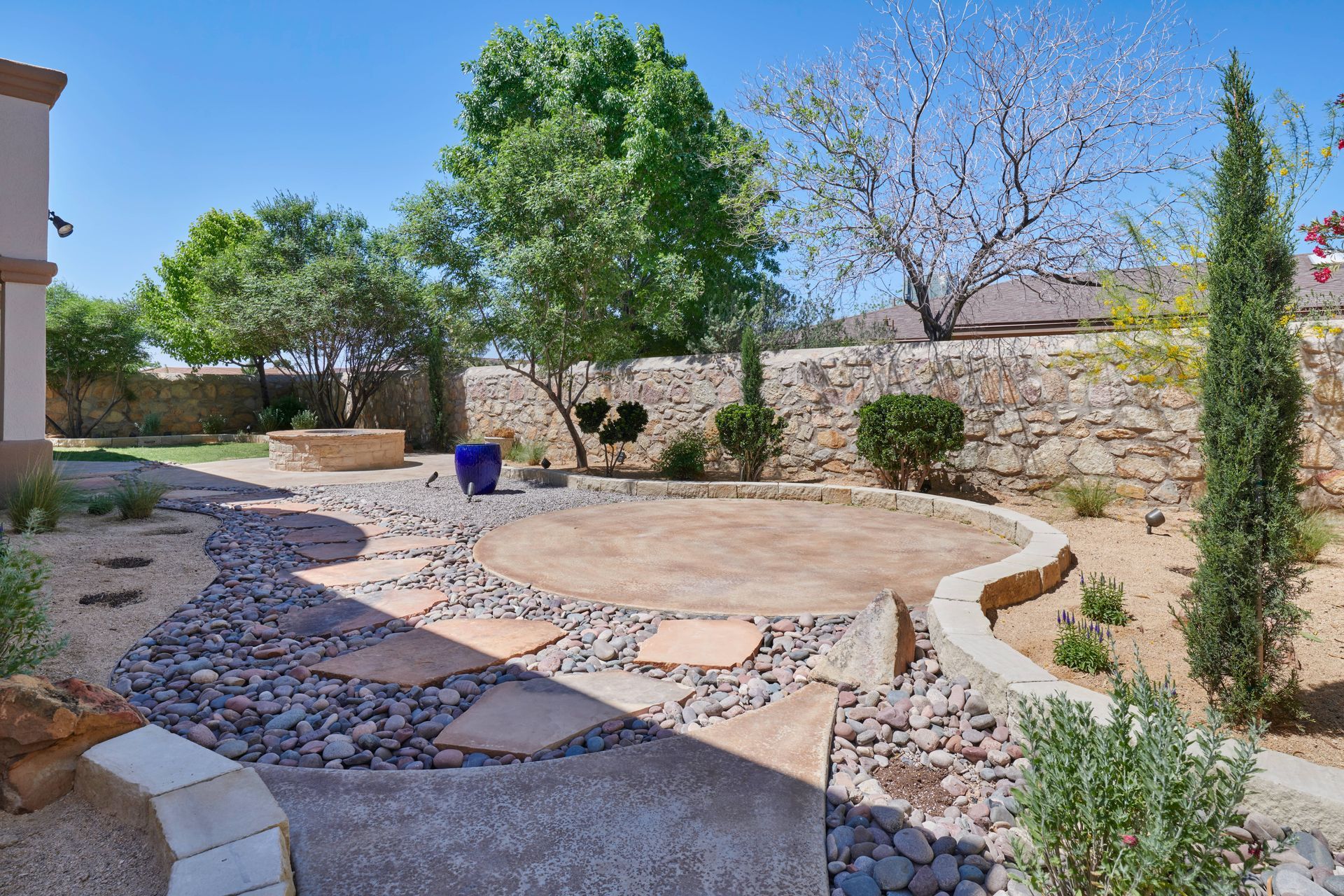 Backyard with circular patio, stone pathway, gravel, trees, and stone wall on a sunny day.