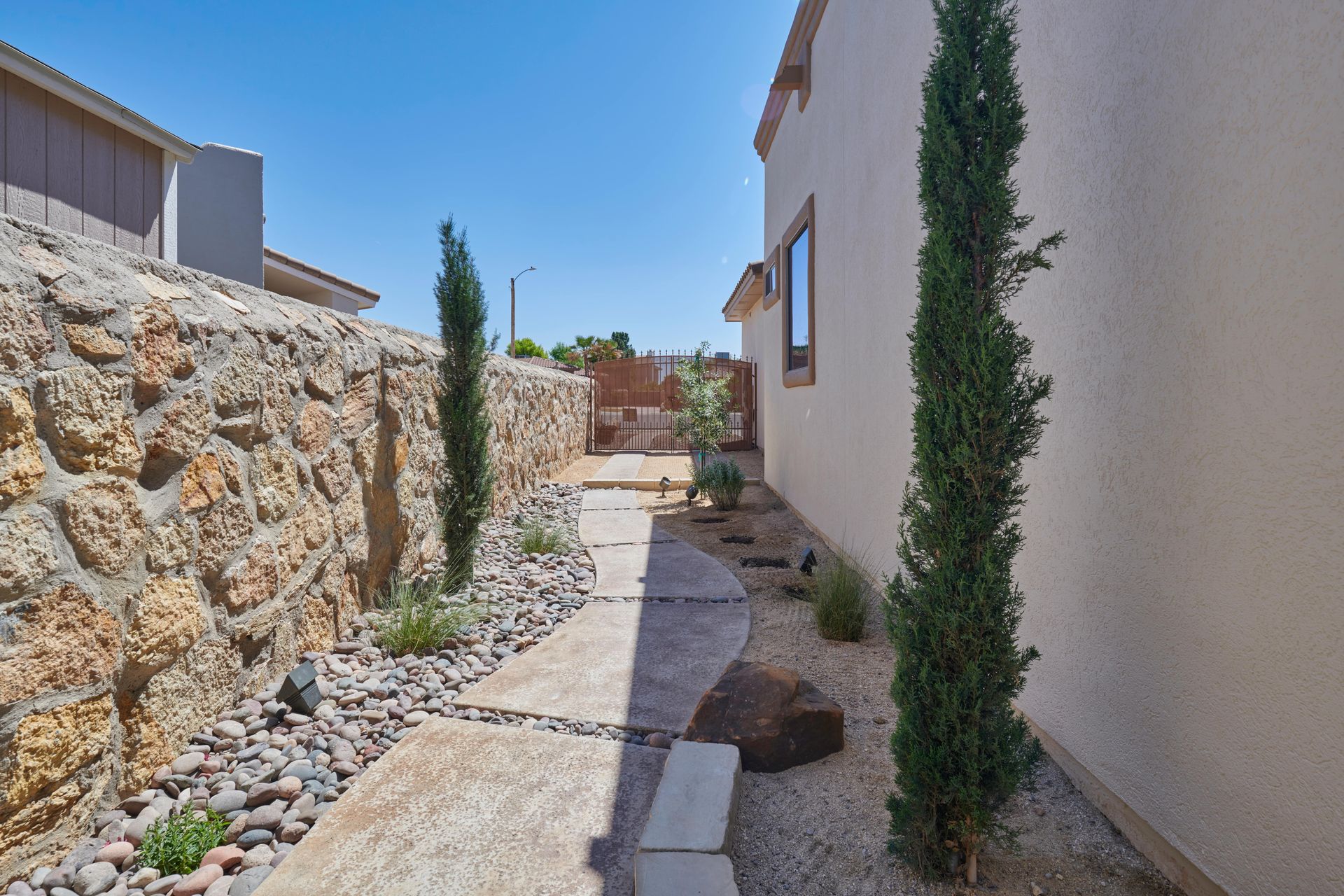 Stone pathway between a stone wall and a white building with cypress trees and landscaping.