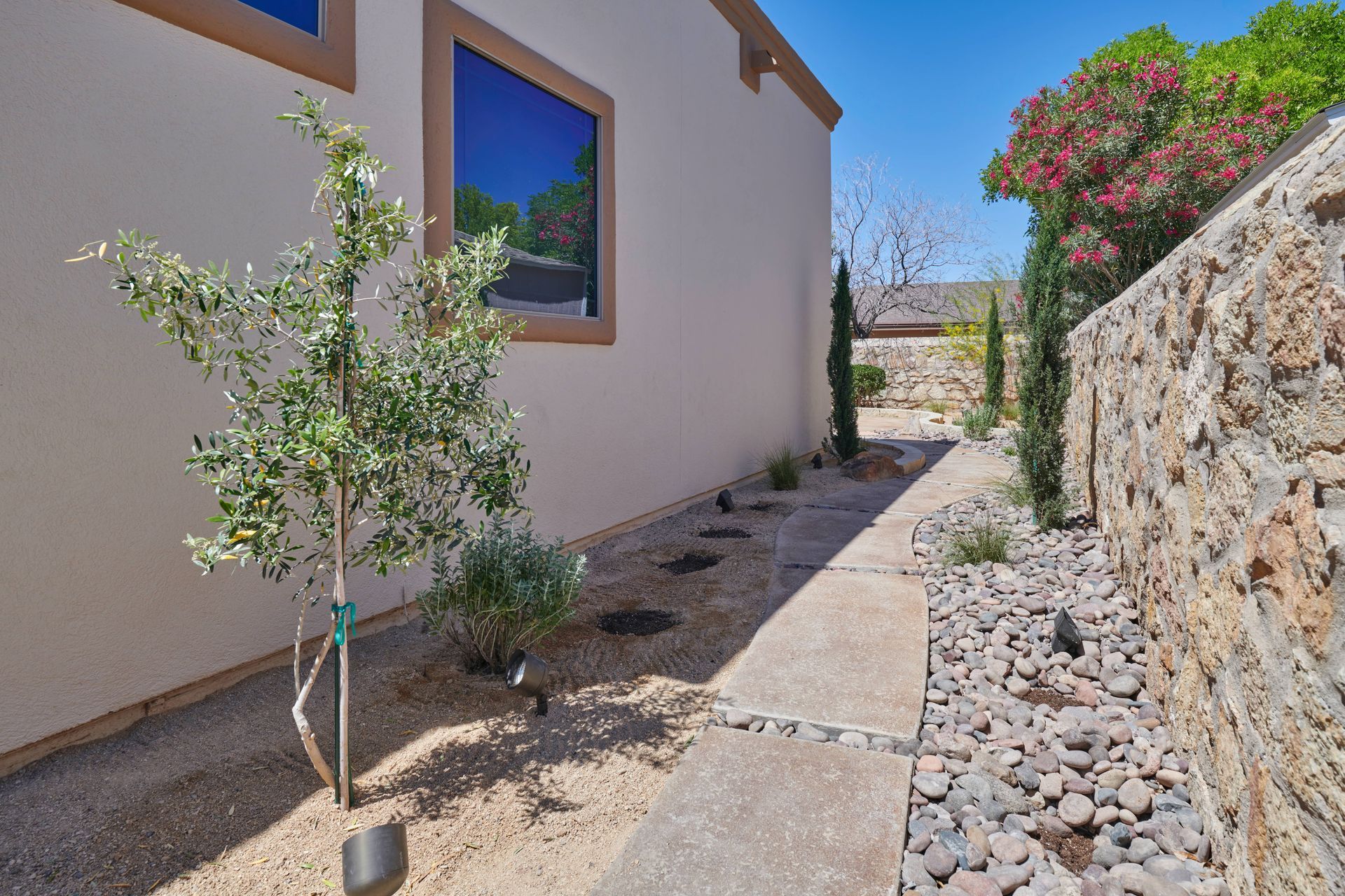 Exterior pathway with a stone wall, gravel, and small trees next to a light-colored building.