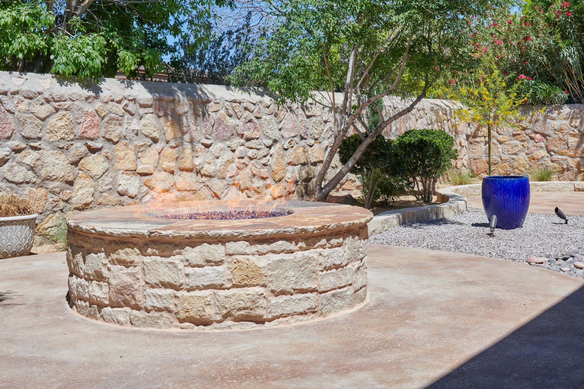 Stone fire pit with flames, in a backyard setting with stone wall and blue vase.