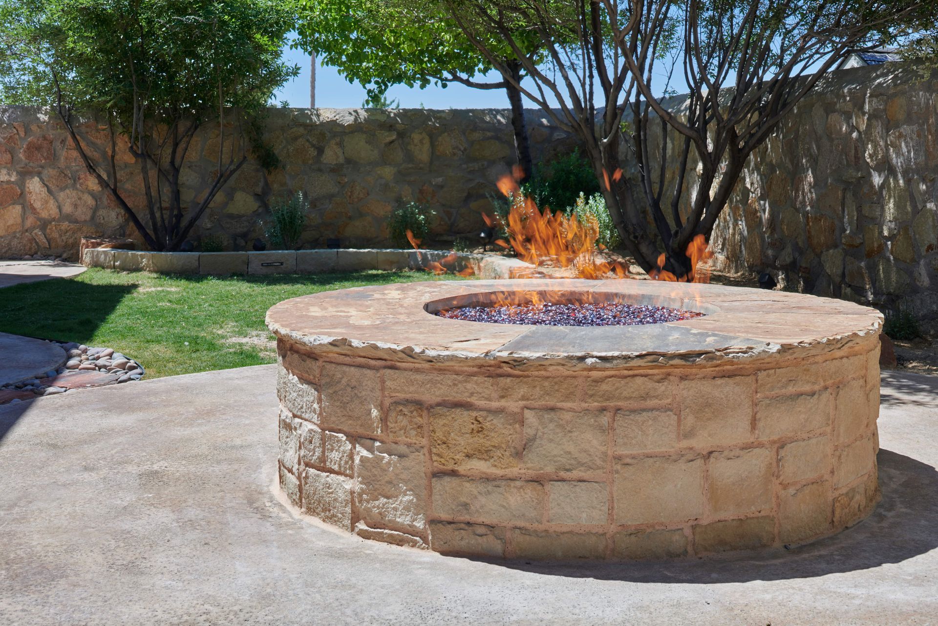 A stone fire pit with flames, in a backyard setting with trees and a stone wall.