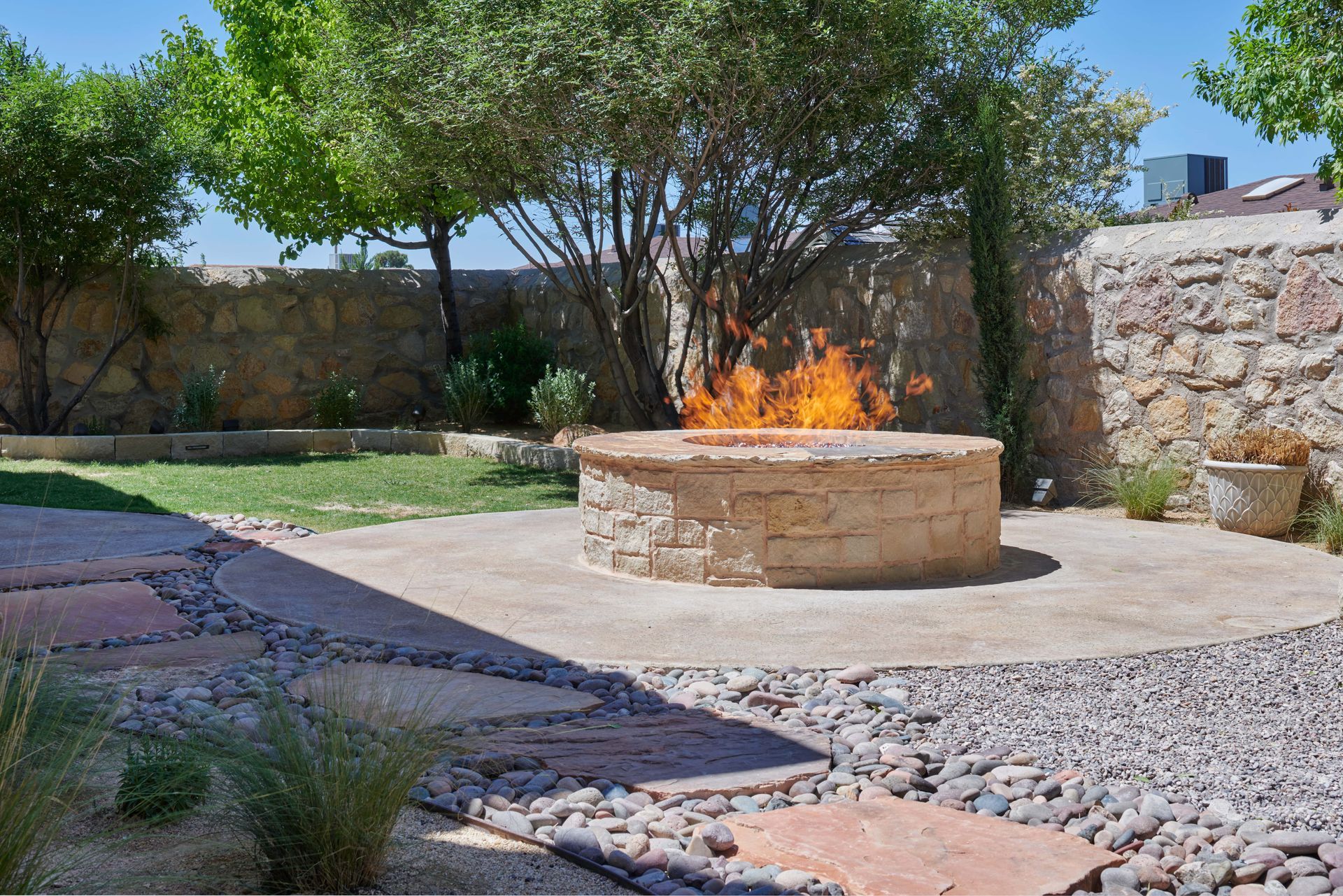 Stone fire pit with flames, surrounded by circular patio, grass, and stone walls.