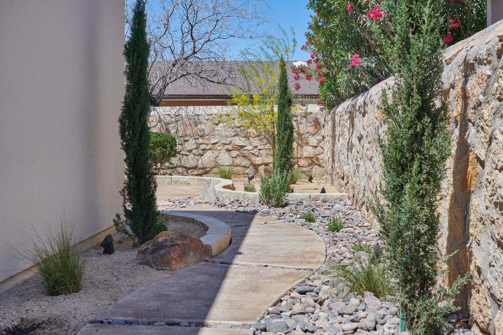 Narrow stone pathway with cypress trees and rock walls, leading to a sunny garden.