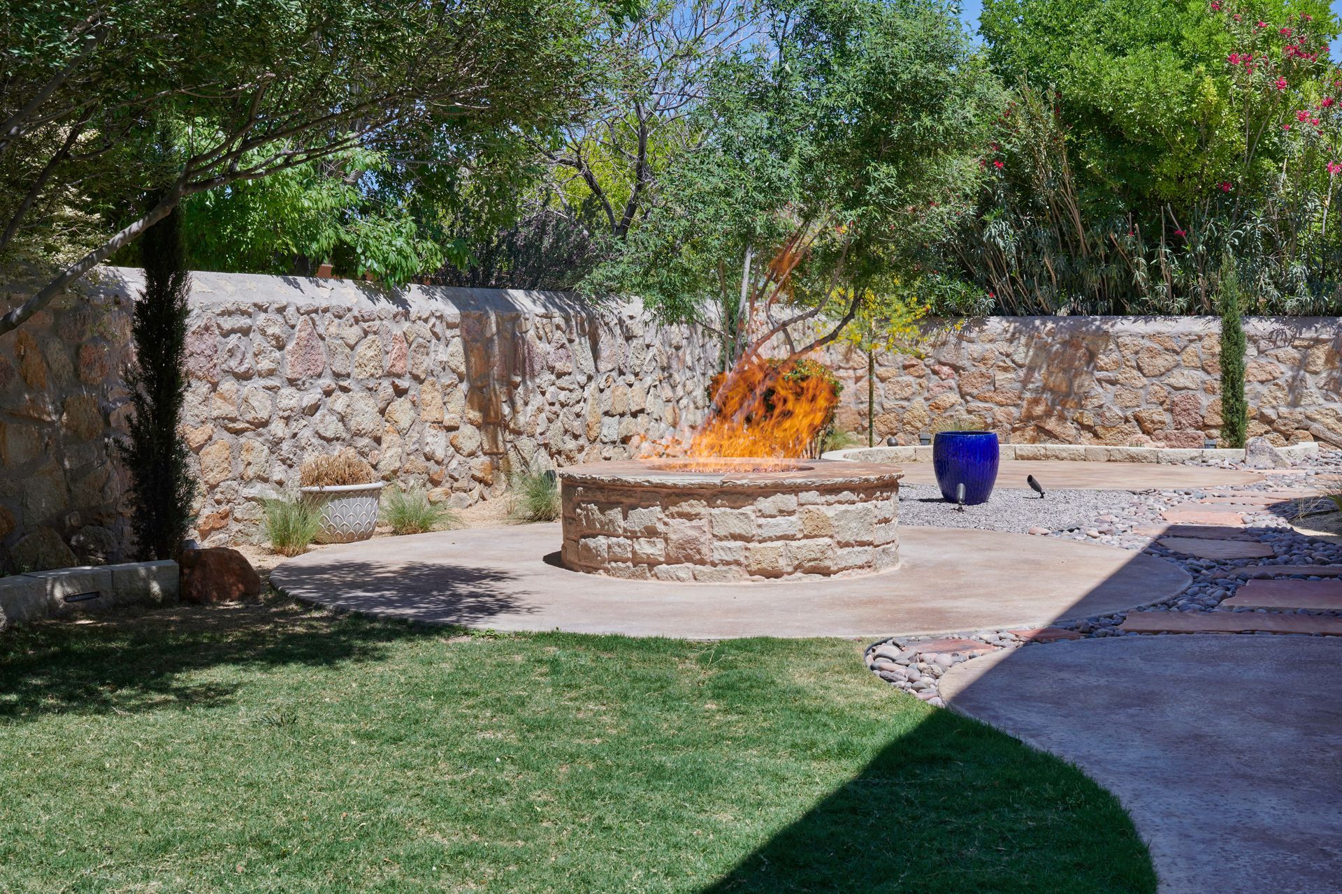 Stone fire pit with flames, in a backyard with green grass and stone wall.