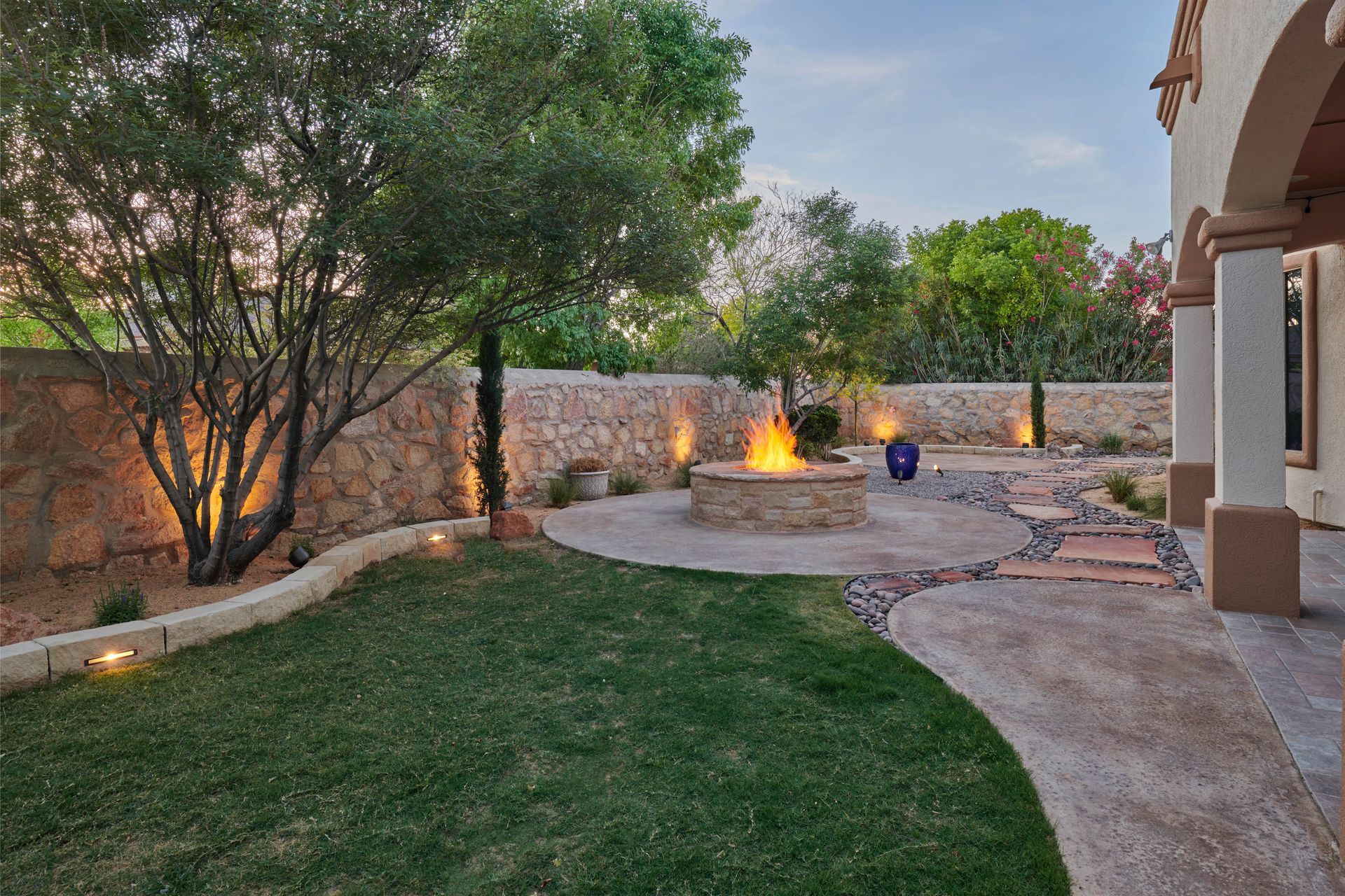 Backyard with fire pit, stone wall, green grass, trees, and pathway lit with soft lighting.