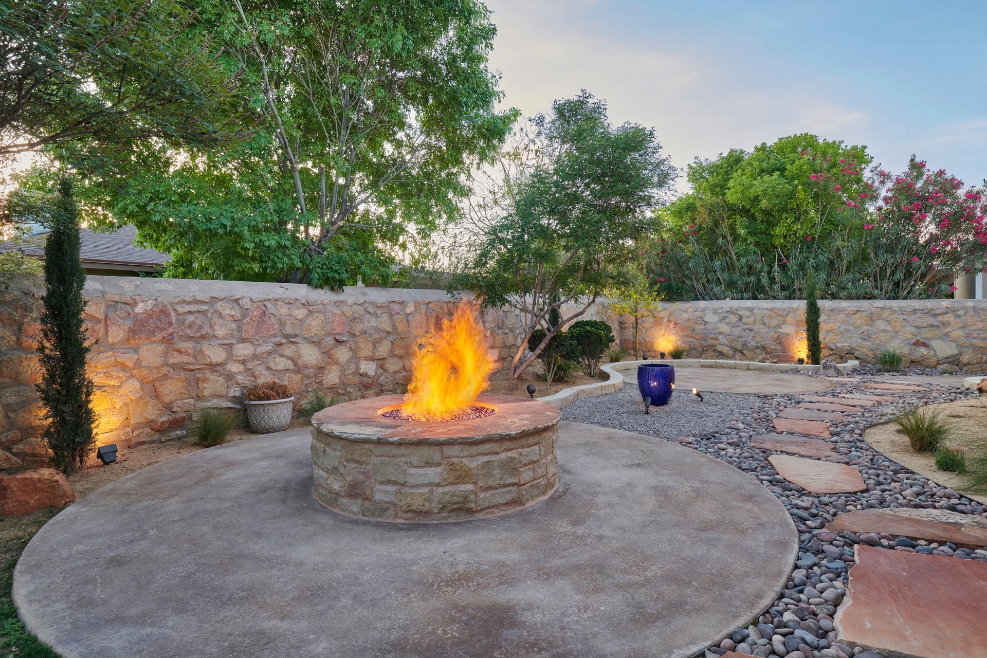 Stone fire pit with flames, on a concrete patio, with a stone wall and landscaping.