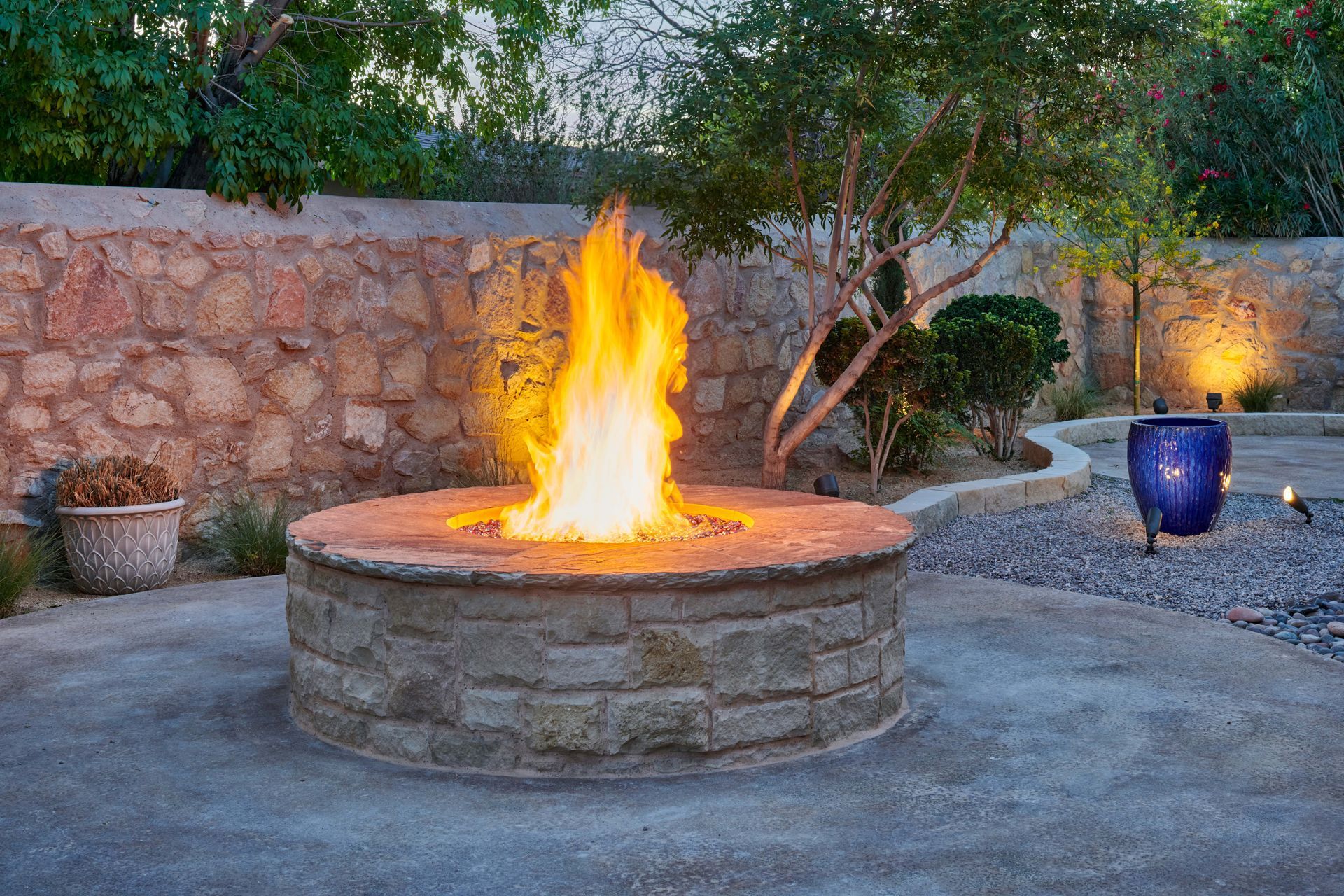 Fire pit with large flames in a stone patio setting, near a wall and shrubbery.
