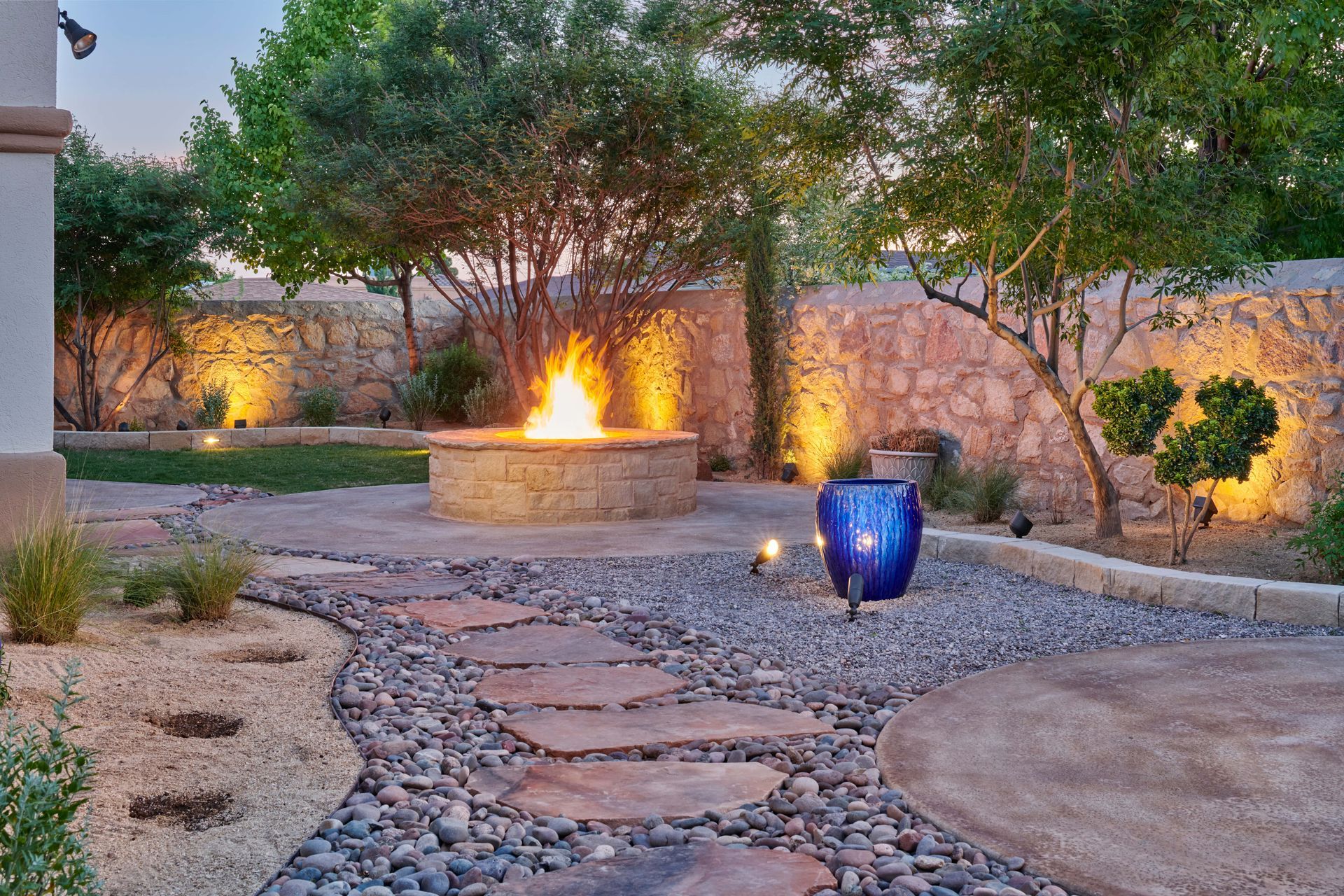 Stone patio with fire pit, pathway, and landscape lighting at dusk.