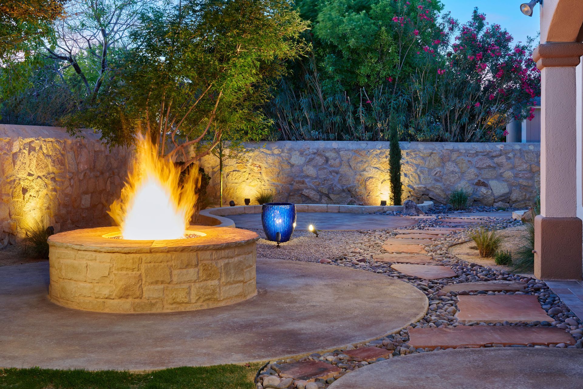A lit fire pit in a stone patio, illuminated by warm lights, with a blue vase and pathway.