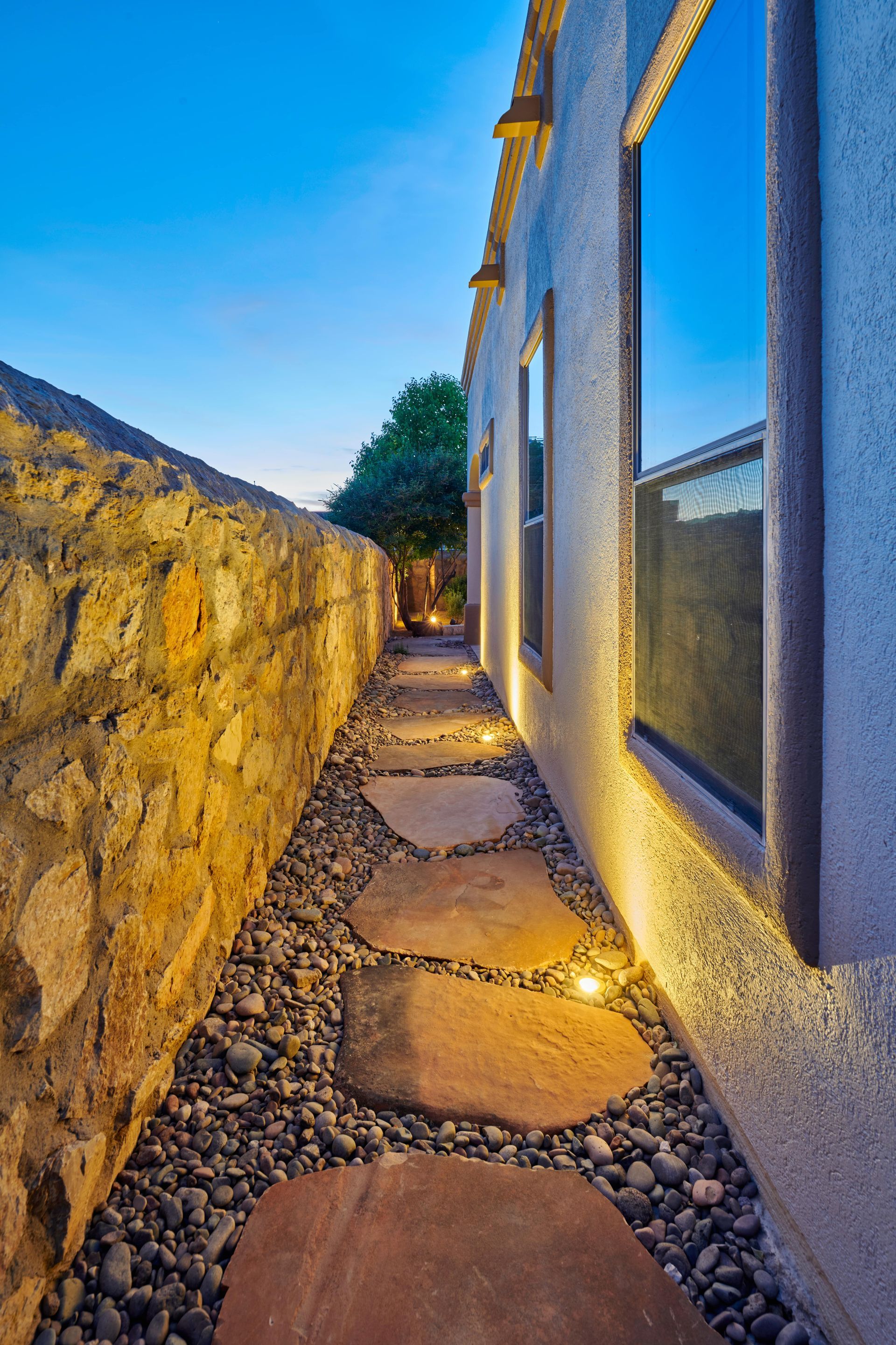 Stone pathway with stepping stones and gravel, flanked by a stone wall and a stucco building with windows, lit at dusk.