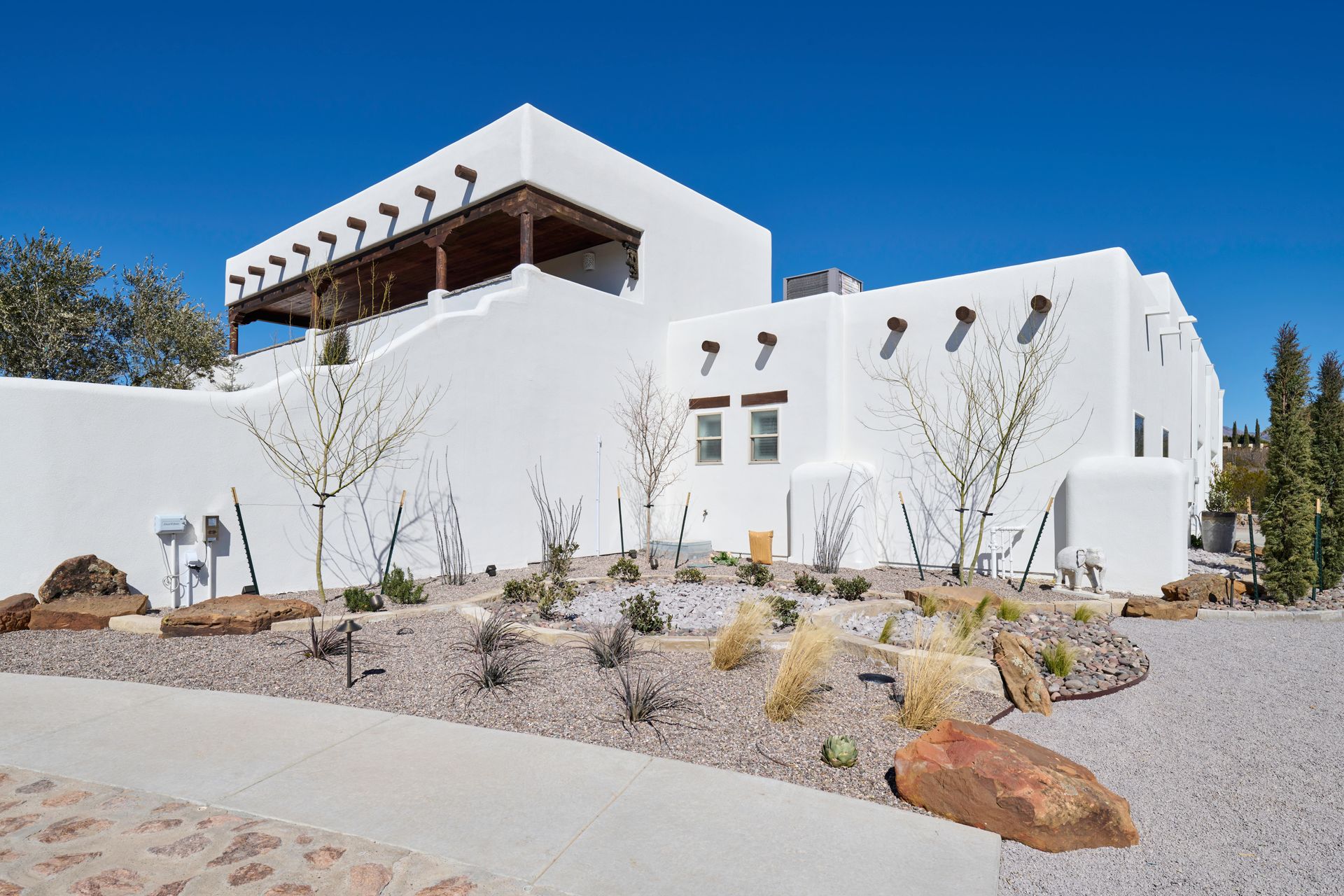 White stucco building with a flat roof, wooden beams, and a landscaped yard under a bright blue sky.