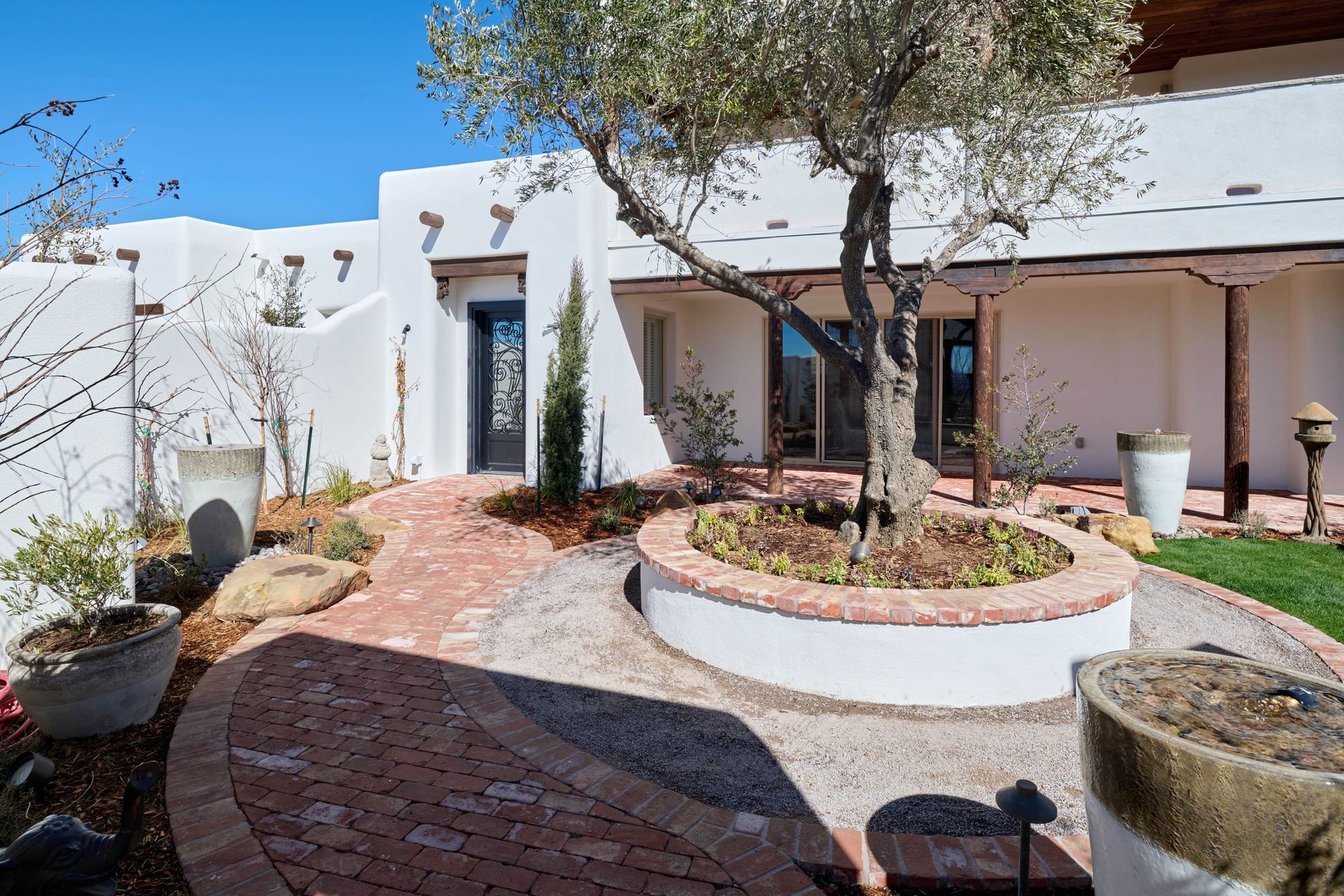 White stucco house with brick pathway, olive tree, and sunny blue sky.