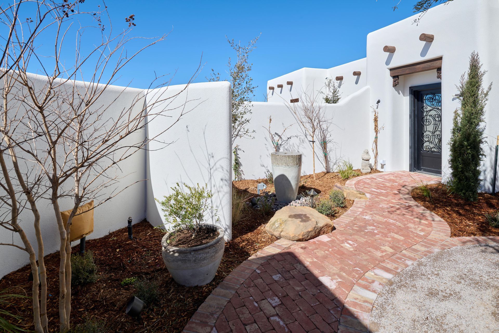Brick path leads to a white stucco building with a dark door, trees, and landscaping.