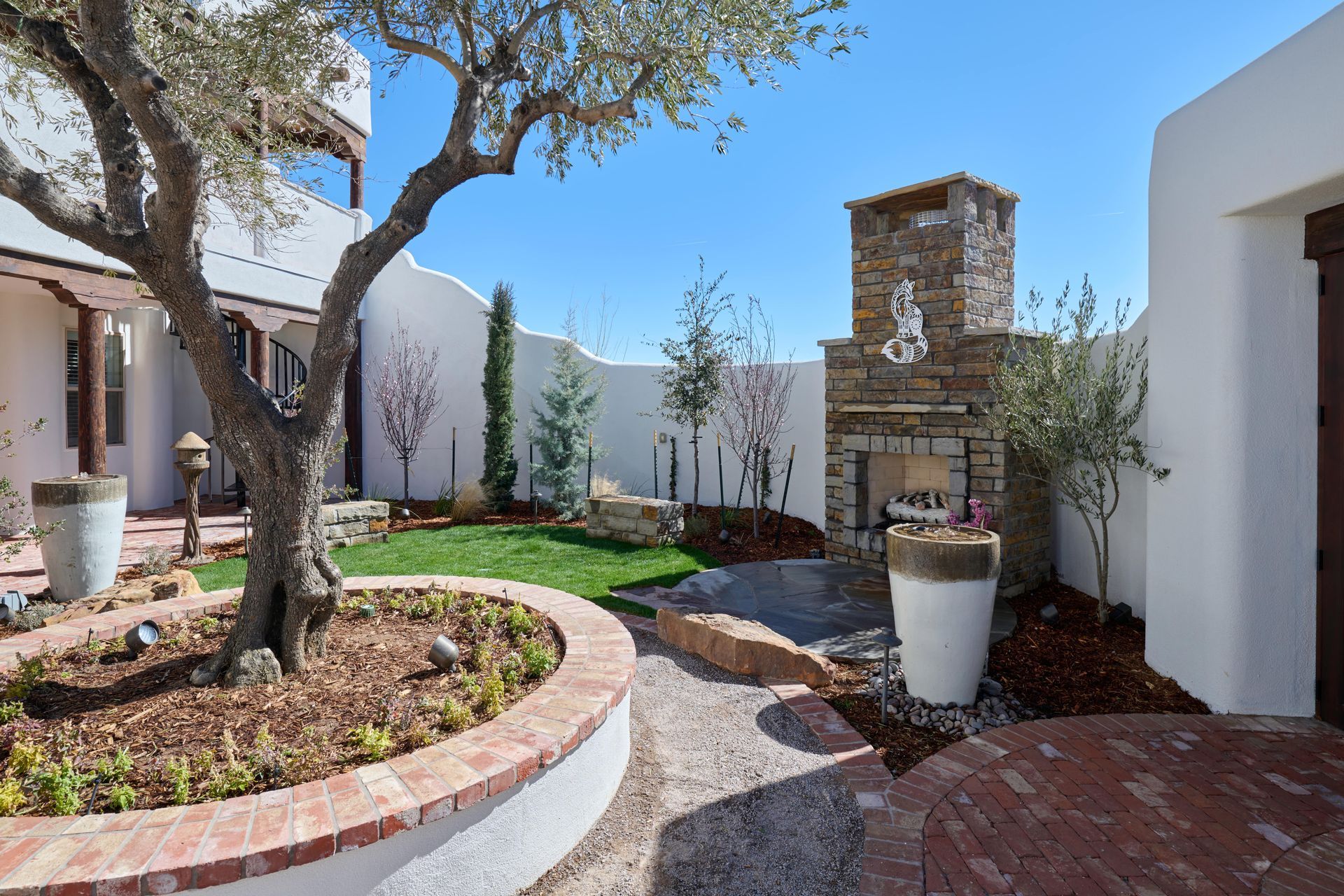 Courtyard with brick patio, fireplace, olive tree, lawn, and white stucco walls. Bright sunny day.