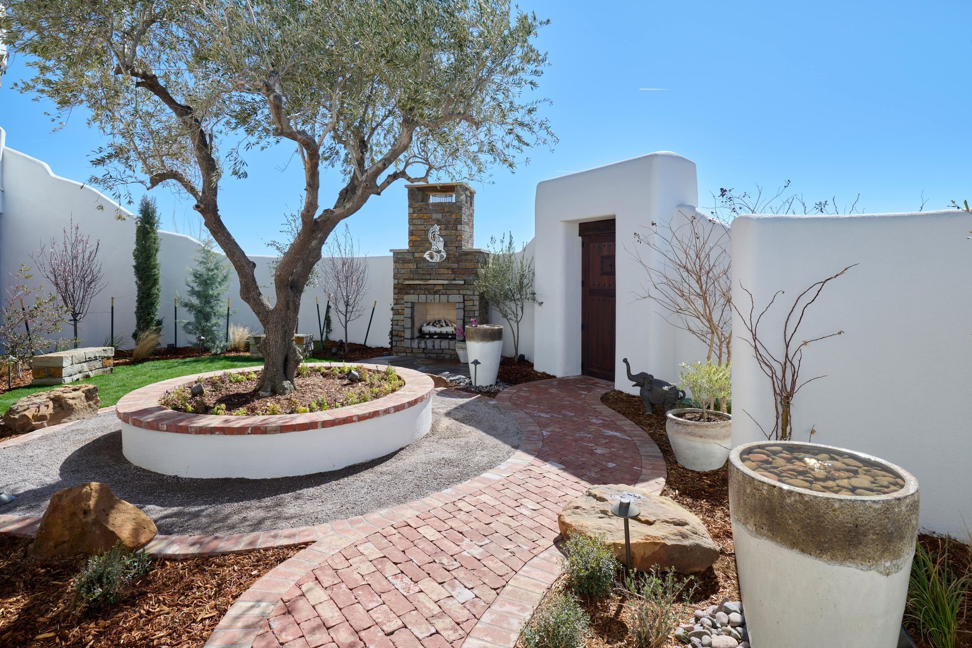 Courtyard with olive tree, brick path, fireplace, and white walls under blue sky.