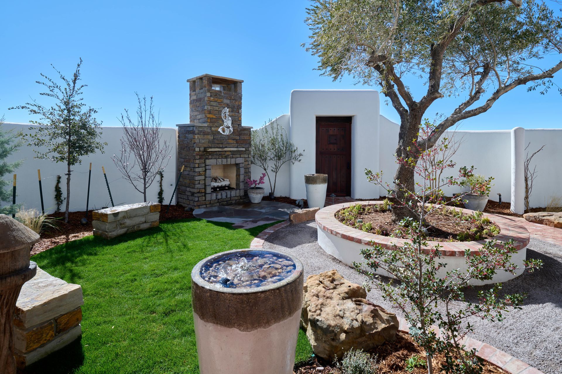 Outdoor patio with fireplace, mature olive tree, and water feature on a sunny day.