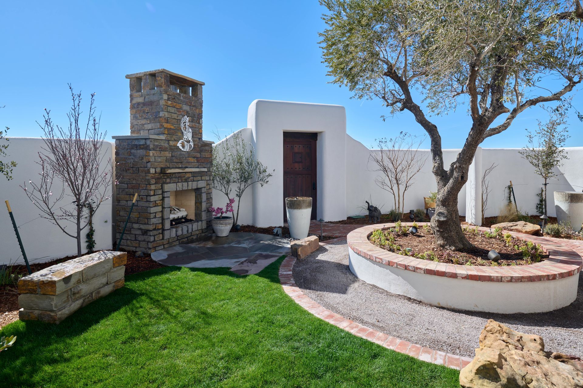 Outdoor patio with fireplace, mature olive tree, white walls, and green lawn.