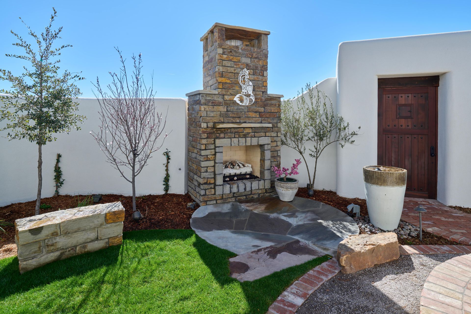 Stone outdoor fireplace on a patio, surrounded by green grass, small trees, and a white stucco wall.
