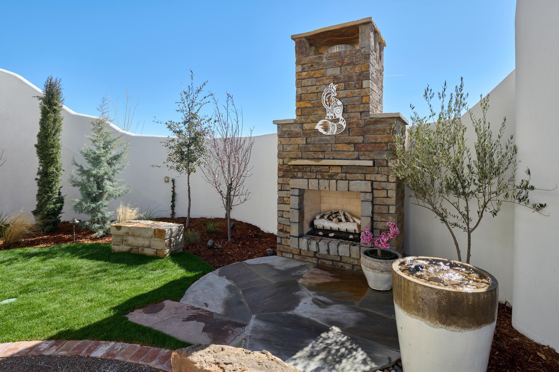 Outdoor stone fireplace in a courtyard with a small lawn, trees, and a decorative planter against a white wall.