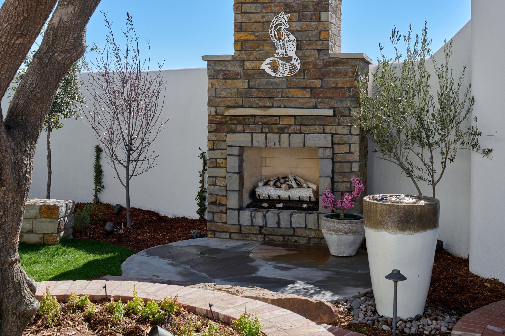 Outdoor stone fireplace with seating area, trees, and white wall.