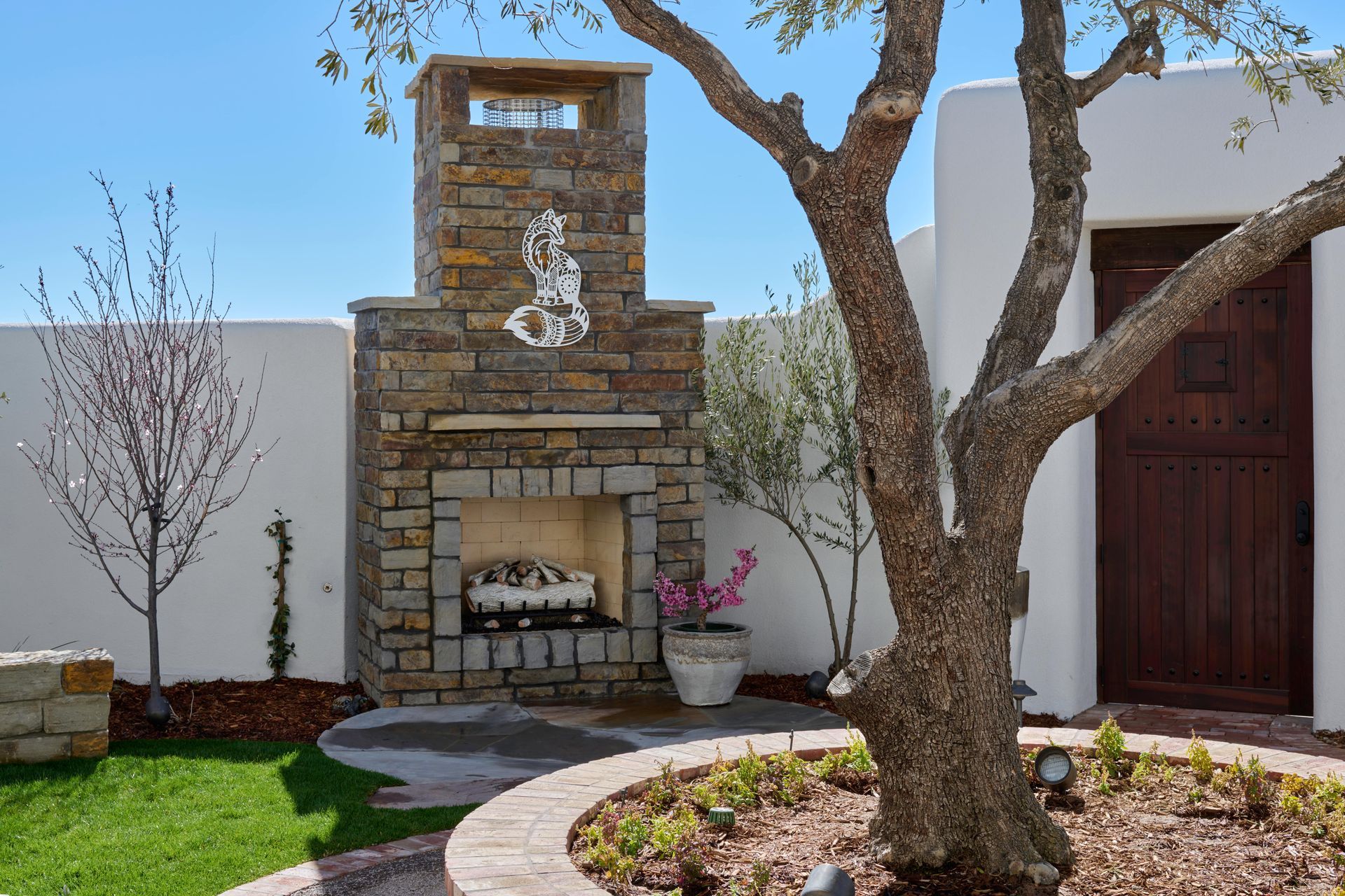 Outdoor stone fireplace with a tree and white walls on a sunny day.