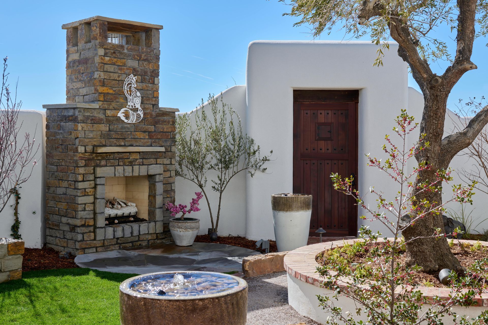 Outdoor patio with a stone fireplace, fountain, and mature tree against a white wall and blue sky.