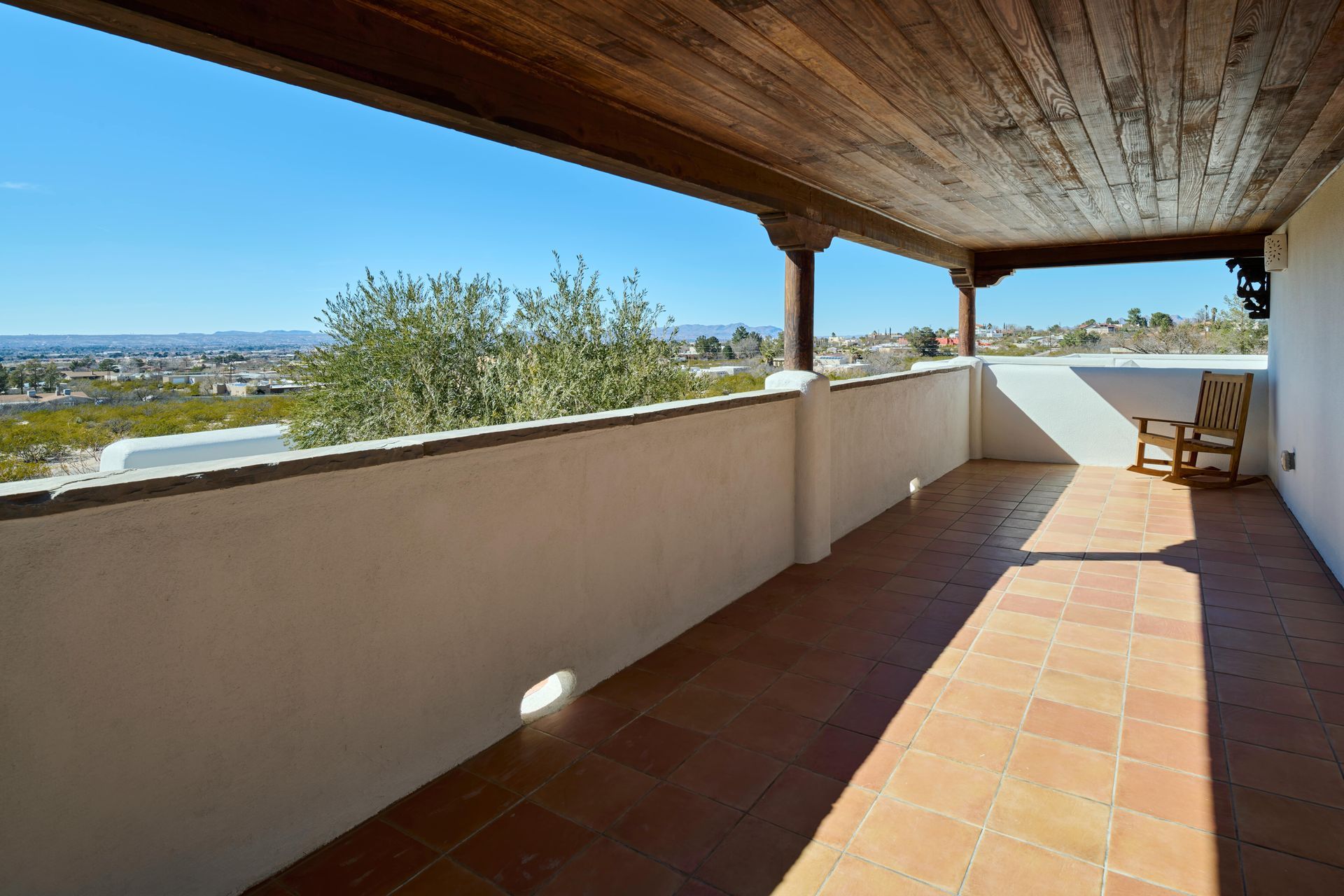 Terracotta-tiled balcony with a wooden ceiling, overlooking a distant cityscape under a bright blue sky.