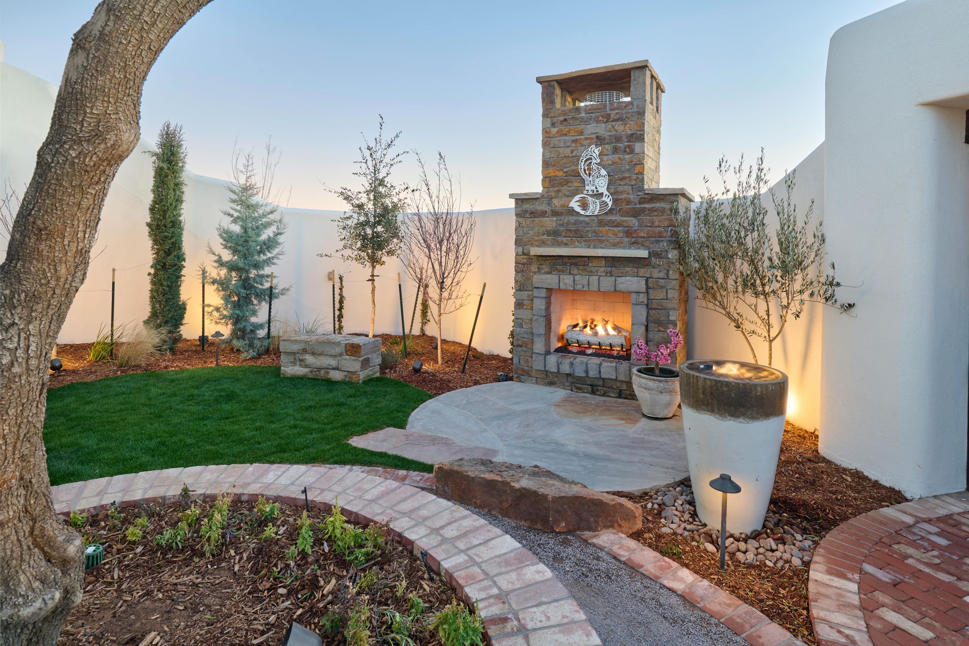 Outdoor fireplace with stone facade in a landscaped yard at dusk. Green grass, brick path, and stucco walls.