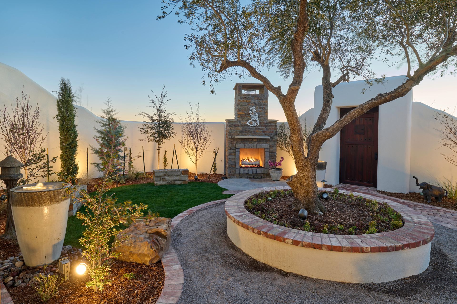Outdoor space with fireplace, mature tree, curved gravel path, and stucco walls.